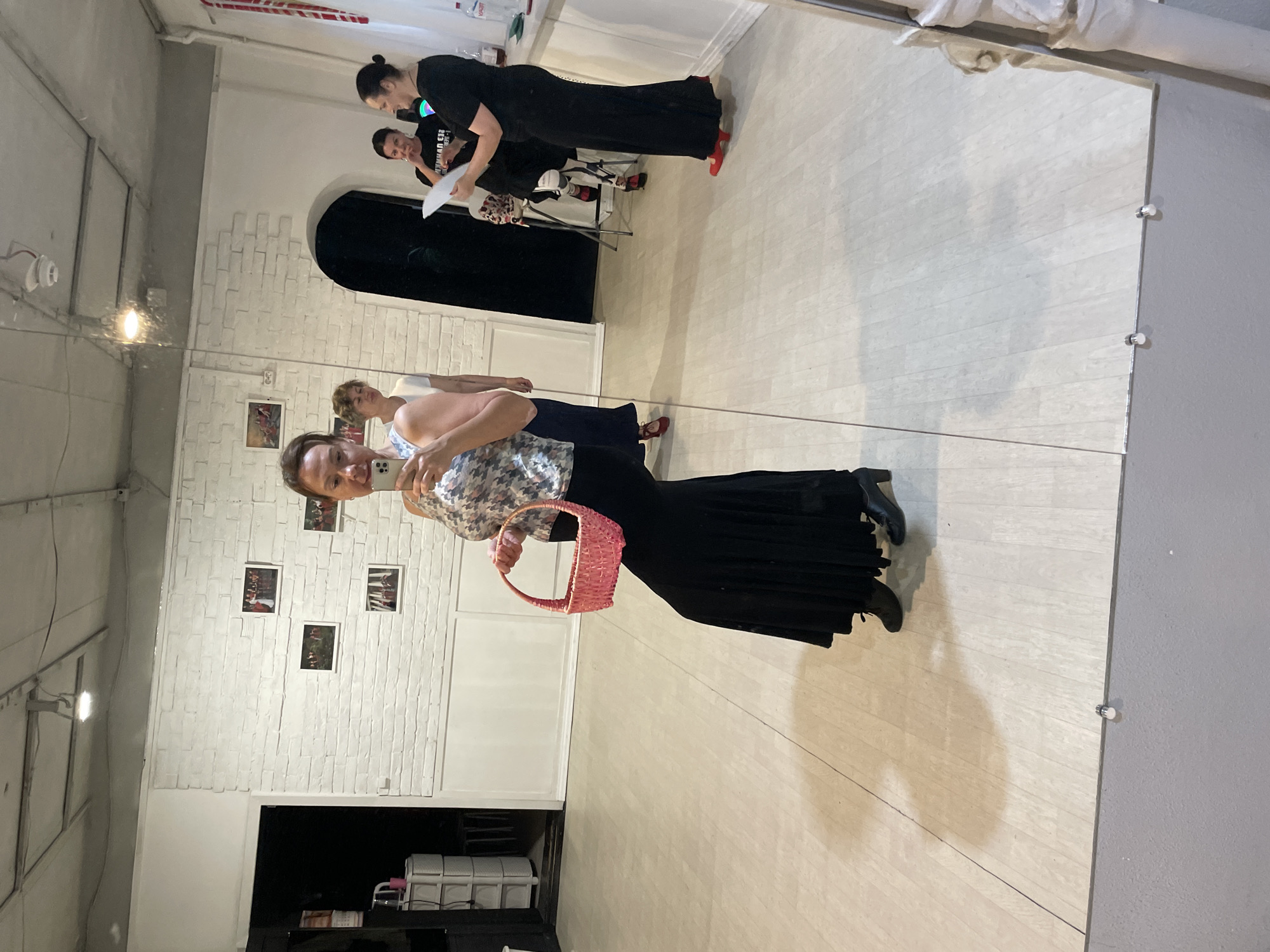 The image shows a woman in a long black skirt and a patterned top, holding a pink basket and a mobile phone in her hand, taking a selfie in a dance studio. She is wearing black boots. In the background, other women are visible, some standing and one sitting, with papers in their hands. The studio has white brick walls with framed pictures and a light-colored wooden floor. The scene appears to be in Sochi, Russia, possibly at a dance or event venue.
