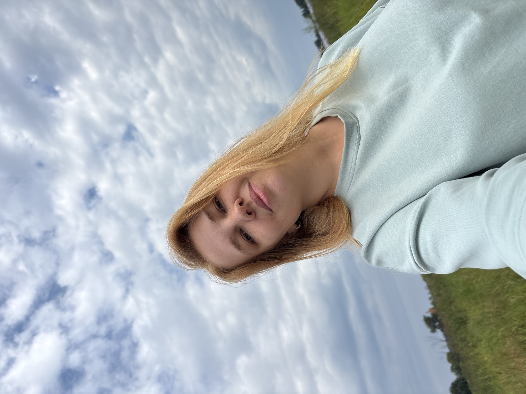 A woman with blonde hair is taking a selfie outdoors. She is wearing a light blue sweatshirt. The background shows a grassy field under a cloudy sky. The location is Breytovo, Russia.