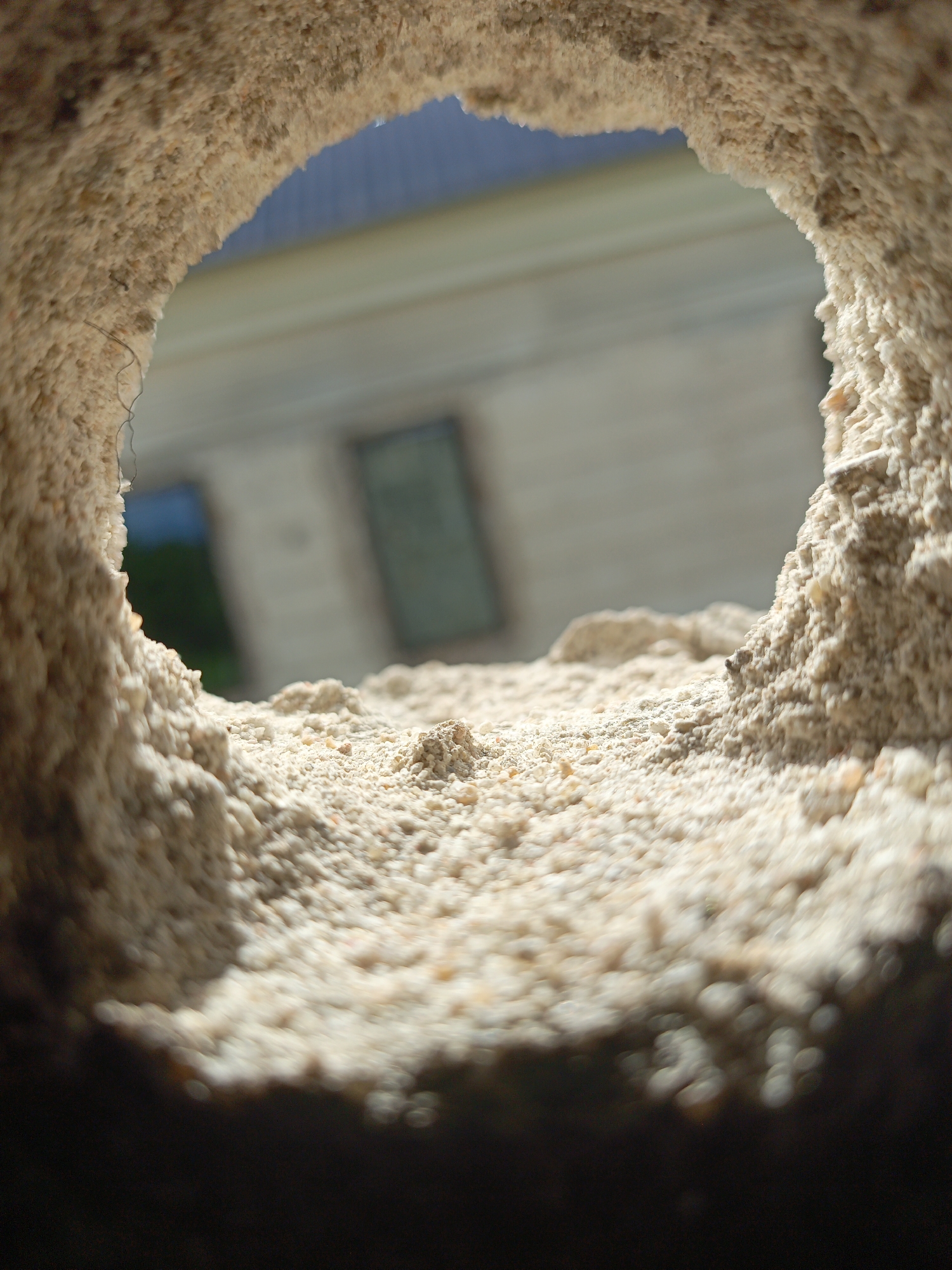 View through a hole in a light-colored, granular material, possibly a wall or rock formation, revealing a blurred building with windows in the background. The foreground material is illuminated by sunlight. The exact location is unknown.
