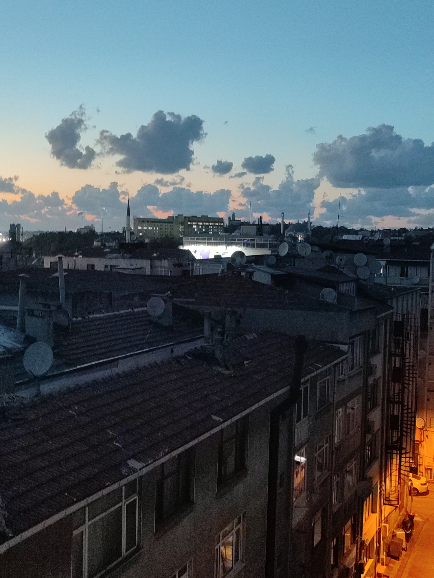 This image depicts a twilight cityscape, likely in Kadıköy, Turkey, as suggested by the architectural style and common urban elements. The sky transitions from a soft orange glow near the horizon to a deep blue overhead, with scattered, dark clouds. In the foreground, a dense cluster of buildings with tiled roofs dominates the scene. Many of these structures are equipped with numerous satellite dishes, indicating a common feature of urban living in this region.
The buildings appear to be residential or mixed-use, with visible windows and some architectural details like chimneys and air conditioning units. Towards the background, larger buildings are visible, including what appears to be a prominent mosque with a minaret, a common landmark in Turkish cities. There is also a larger, more modern-looking building with many illuminated windows, possibly an office building or hotel. The street level, visible on the right side of the image, is illuminated by warm-toned streetlights, casting an orange glow on the pavement and the side of a building. A yellow car is parked on the street, and there are a few bicycles and what look like storage units or air conditioning units against the building wall. No people are clearly visible in the frame, creating a sense of quietude. The overall impression is of a densely populated urban environment during the transition from day to night.
