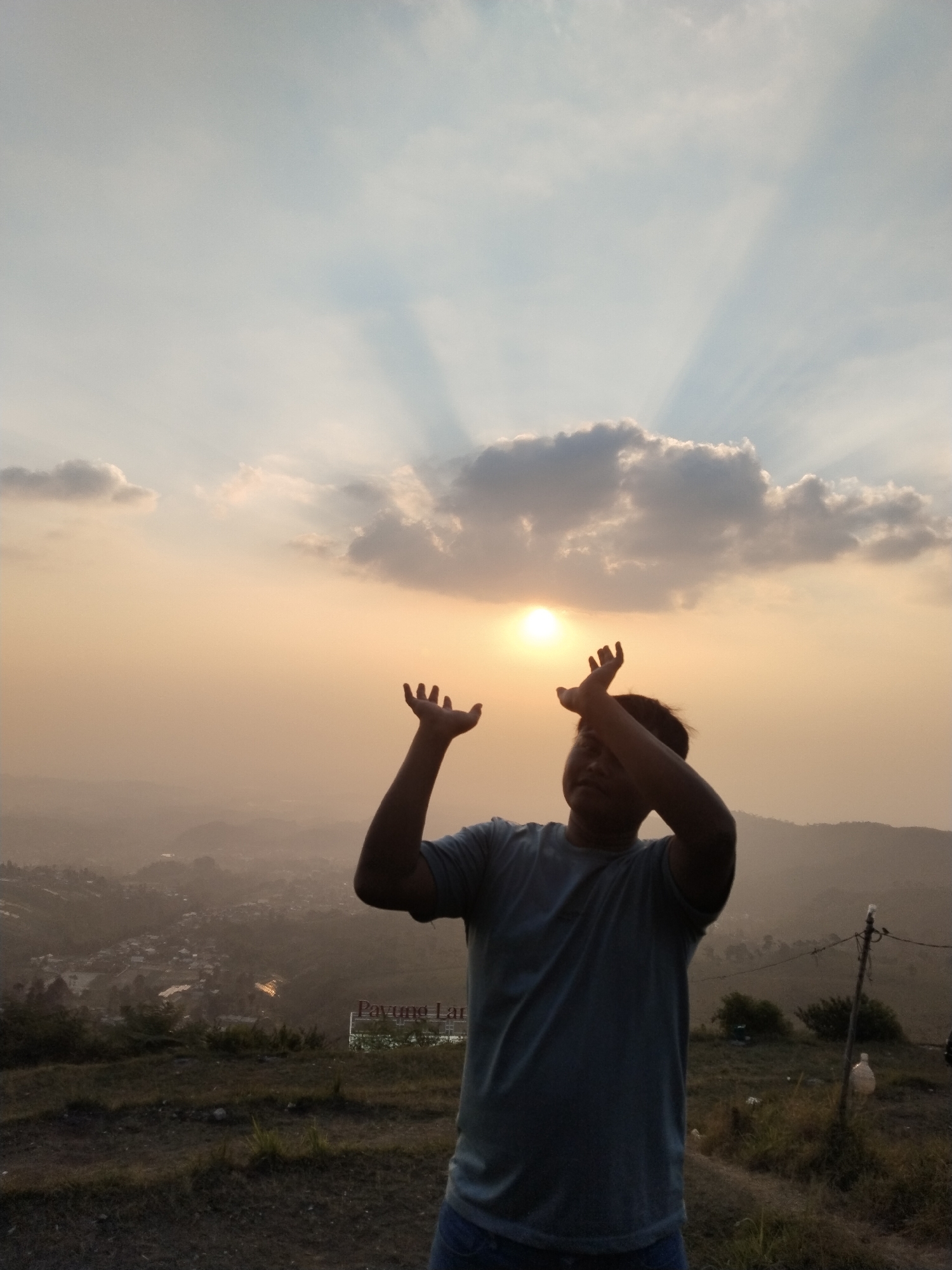 A young man stands on a grassy hilltop, silhouetted against a hazy sunset in Gumeng, Indonesia. He wears a blue t-shirt and jeans and has his arms raised, palms facing upwards, as if reaching for the sky. The sun, partially obscured by clouds, casts long rays of light through the atmosphere. Below him, a valley unfolds with a small town scattered across the landscape, some lights visible. A signpost reading "Payung Lar" is visible in the middle ground. The overall impression is one of awe and tranquility.