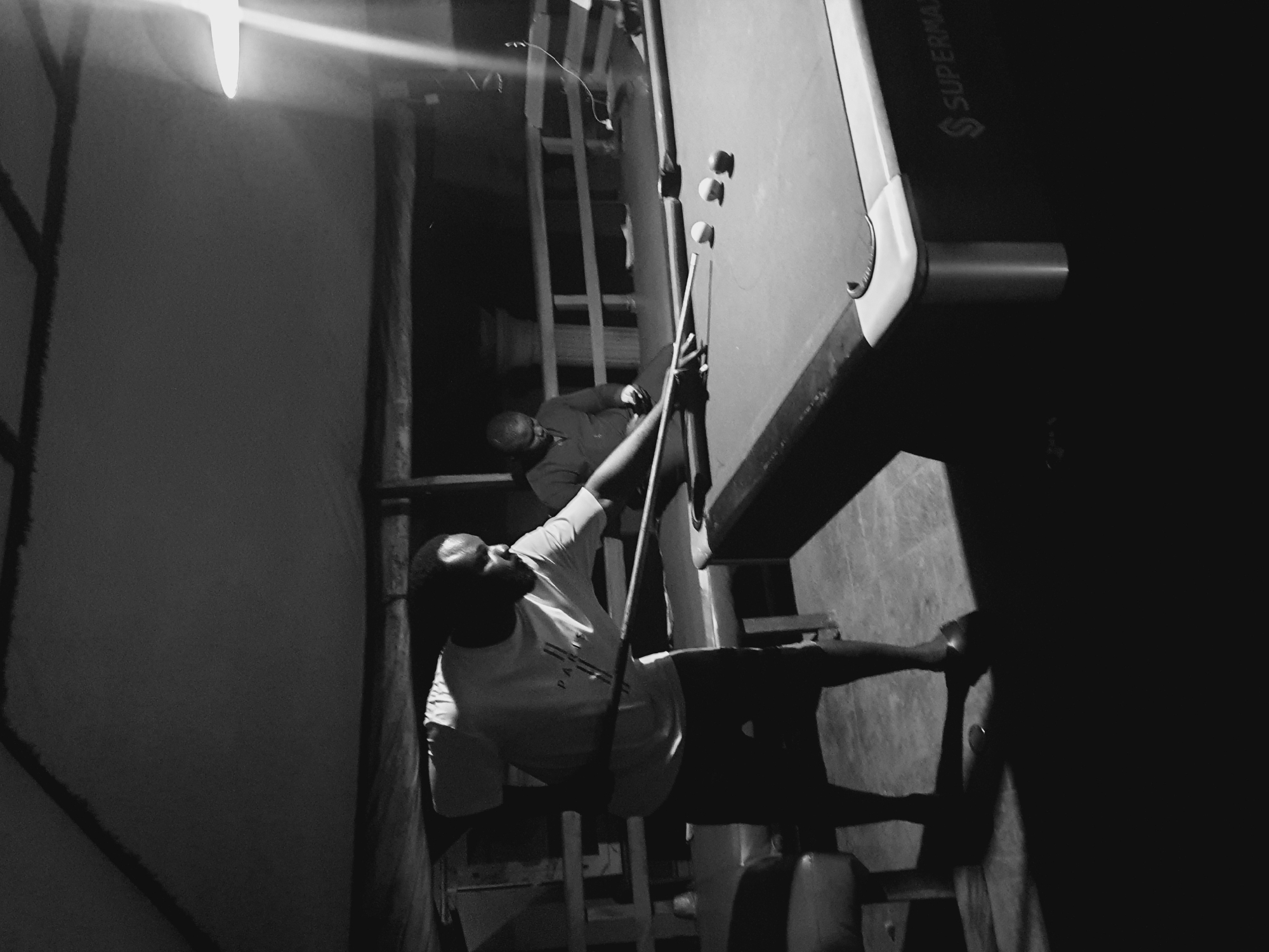Two adult males are playing pool in an indoor, dimly lit space, likely a recreational area in Lokoja, Nigeria. The man in the foreground, wearing a t-shirt and shorts, is in mid-action, aiming a pool cue at a ball on a dark green pool table. He has a beard and short hair. The second man, dressed in a shirt, is visible in the background, partially obscured, appearing to be observing the game. The environment is rustic, with exposed ceiling beams and wooden railings. A bright light source, possibly a lamp, illuminates the scene from the right, casting long shadows. The pool table has several balls on it, and the brand "SUPERMAX" is visible on its side. The flooring appears to be concrete.