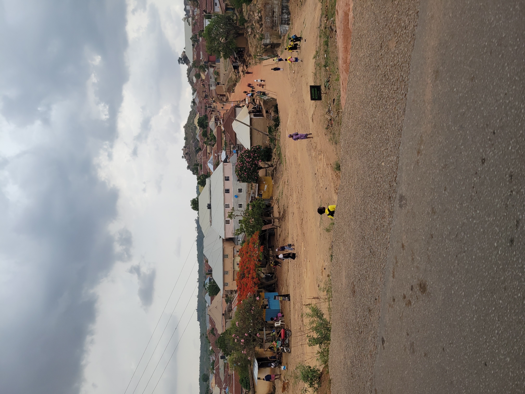 A slightly rotated medium shot shows a dirt road that runs alongside an asphalt road. The image depicts a densely populated area, possibly a suburb or a neighborhood on the edge of the city. The architecture is varied, ranging from modest single-story structures to taller, multi-story buildings. The area appears to be situated on a hillside.
Several people are visible, engaged in various activities. Some are walking along the dirt road, others appear to be gathered near buildings, and some might be children playing. A sign with text, "FEDERAL SECRETARIAT JOS" can be seen.
The environment features trees and vegetation interspersed among the buildings. The sky is overcast, suggesting it might be a cloudy day. Power lines stretch across the top left corner of the image. The photograph likely depicts a common scene in a residential or commercial area within Jos, Nigeria.
