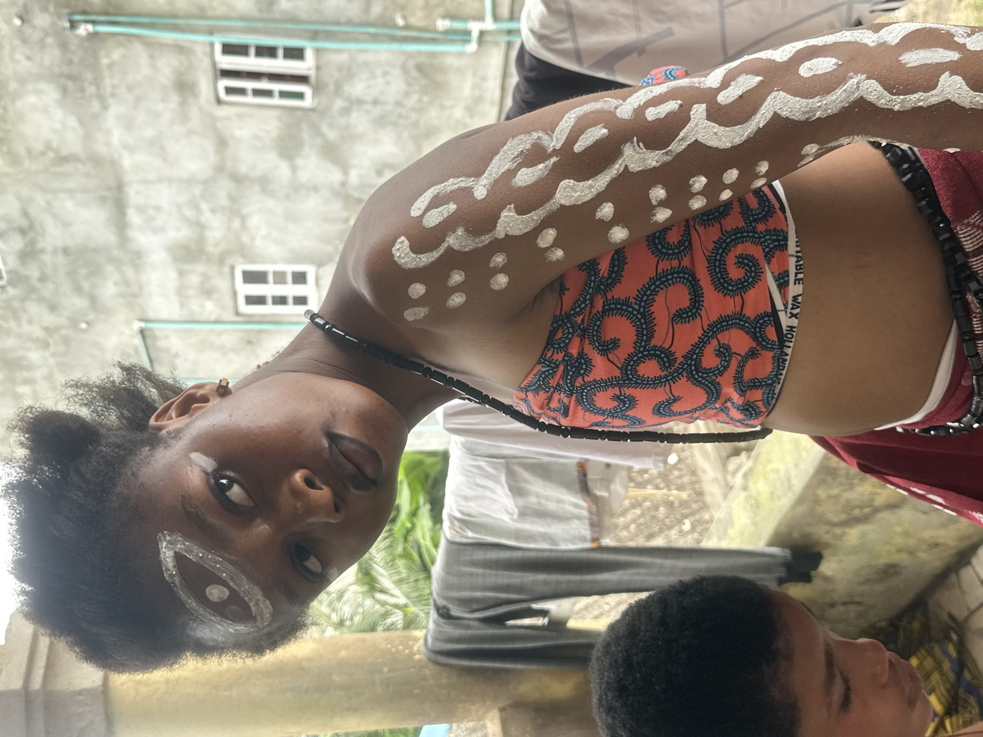 The image shows a close-up of a young Black woman in Badagry, Nigeria. She is wearing a vibrant orange and blue patterned crop top and a red skirt. Her face is turned to the side, and she has white face paint around her eye, resembling an abstract eye design. She is also wearing a black beaded necklace. Her arm is decorated with white body paint, featuring swirling patterns and dots. In the background, there is a building with visible windows, laundry hanging on a clothesline, and lush greenery. To the left, a young Black child's head is visible, looking downwards.