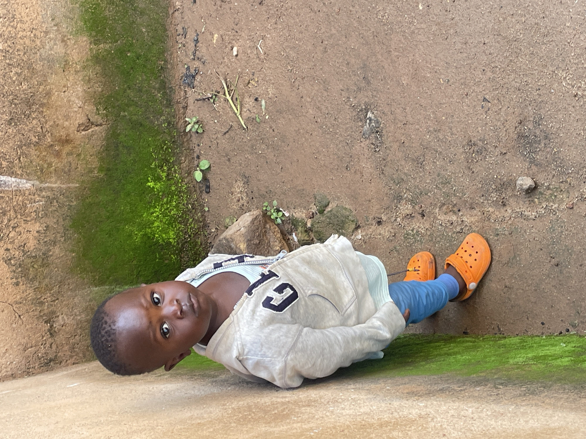 In Barakin Lerawi, Nigeria, a young child stands facing the camera, leaning against a textured, tan-colored wall covered in patches of green moss. The child wears a light gray hooded jacket with a "GAP" logo on the chest, over a striped light blue shirt and blue pants. Bright orange clogs are on the child's feet. The ground is dirt with small pebbles and sparse green vegetation. A large rock is visible to the right of the child, and further beyond is more of the moss-covered wall.