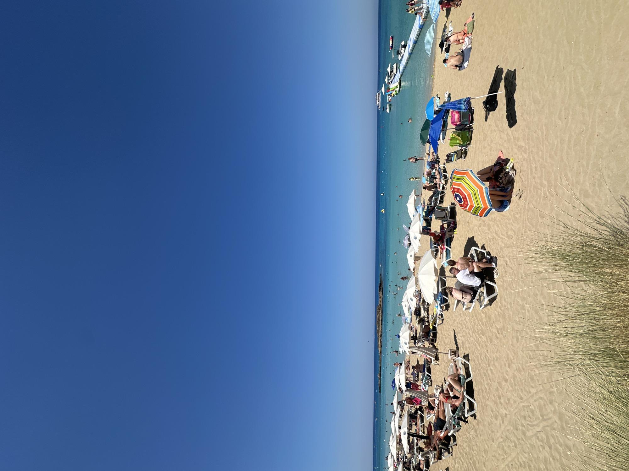This is a wide-angle, eye-level shot of a crowded beach in Protaras, Cyprus, on a clear, sunny day. The foreground is dominated by sand, with a few tufts of dry grass on the left. The middle ground depicts a busy beach scene with numerous people sunbathing on lounge chairs, sitting under umbrellas, and walking around. White beach umbrellas are prevalent, along with a few colorful ones. The background features the turquoise Mediterranean Sea, dotted with several boats and a floating water park with a long pier. The sky is a vibrant, cloudless blue, occupying the upper half of the image.