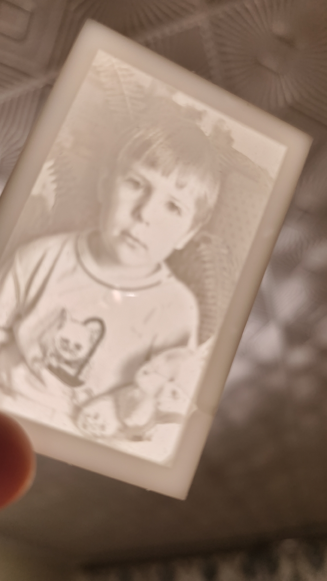 A sepia-toned, faded photograph of a young child, possibly a boy, is displayed in a white frame. The child has a bowl-cut hairstyle and is wearing a long-sleeved shirt with a graphic of a cat on it. They are holding what appears to be a stuffed animal. The photograph is tilted, and a thumb is visible at the bottom left corner, obscuring a small portion of the frame. The background of the photograph shows a textured pattern. The overall image quality is soft and slightly blurry, suggesting an older photograph or a stylistic choice. The photograph is placed on a surface with a patterned texture, possibly a tablecloth or bedding, which is illuminated from above, casting shadows.