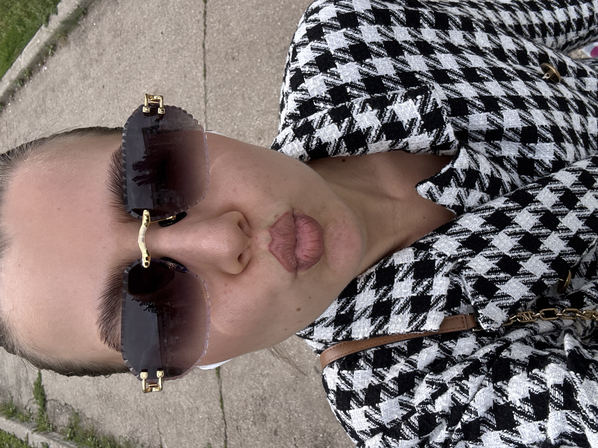 A woman is taking a selfie outdoors in what appears to be a public walkway. She is wearing oversized, rimless sunglasses with a gradient brown tint and gold accents. Her hair is pulled back into a bun, and she is wearing white earbuds. She has on a black and white houndstooth jacket and a brown purse strap is visible over her shoulder. Her lips are pursed in a kiss. The background shows a concrete path and some grass.