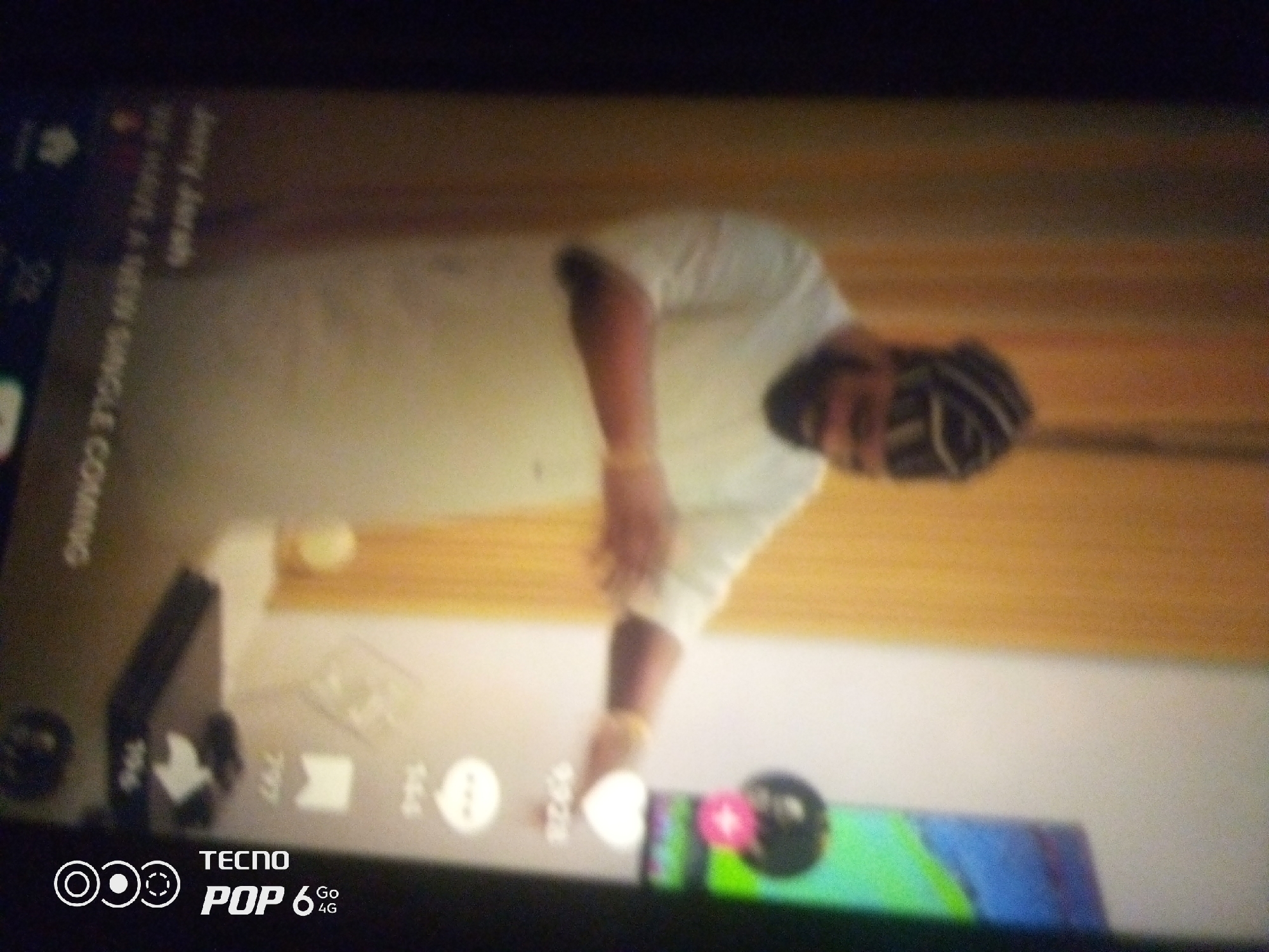The image is a low-resolution, blurry, and rotated photograph taken with a mobile phone, likely a Tecno POP 6 4G, as indicated by the logo at the bottom. It depicts a person wearing a white shirt and a headscarf, standing in what appears to be an indoor setting. The background consists of a wooden surface and a lighter colored wall. There are some user interface elements visible on the screen, including icons and text, suggesting the image was captured from a screen or is a screenshot. The person's posture and the context of the screen elements hint at the possibility of a video or social media platform being displayed. There is no discernible information about the specific location in the city of Howayi, Nigeria.