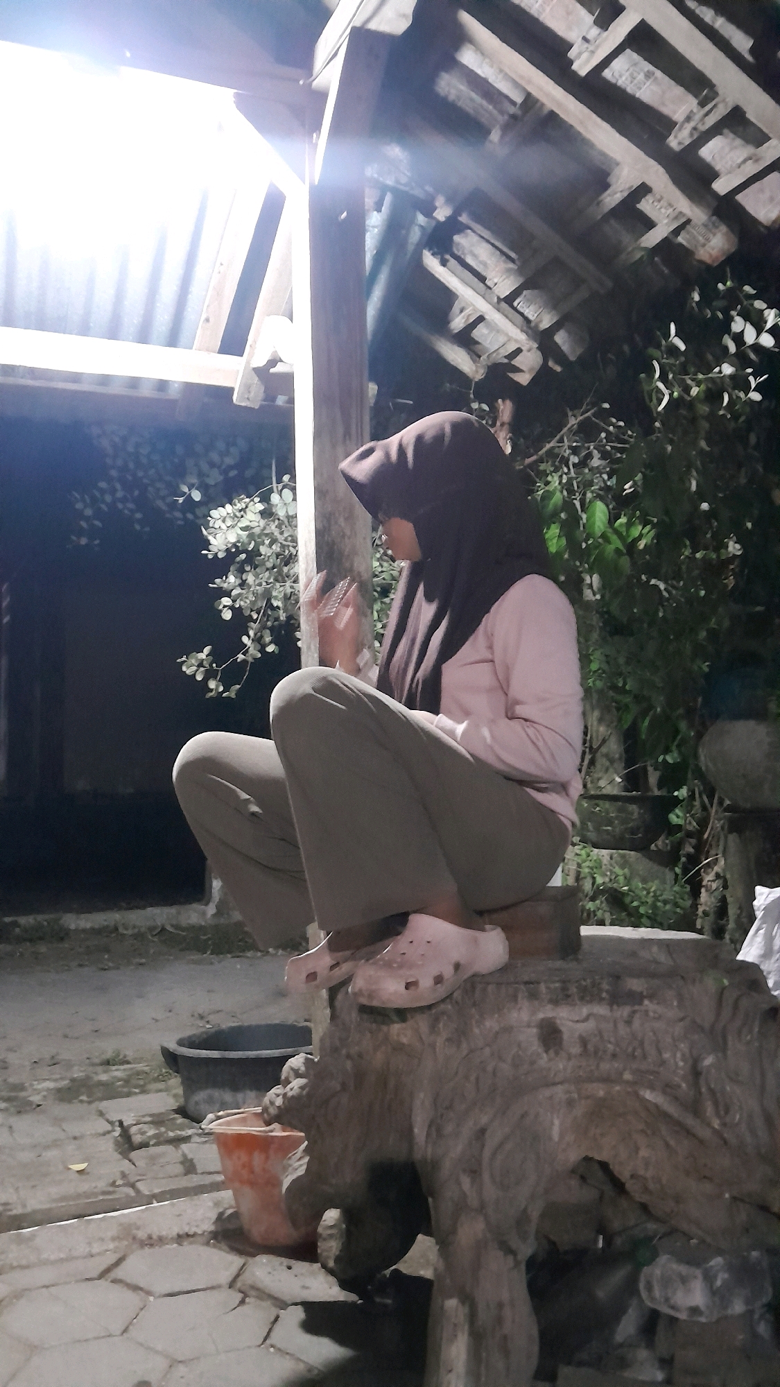 A person wearing a dark brown hijab and a light pink long-sleeved shirt is sitting on a carved stone object in an outdoor setting at night. The person is also wearing light gray pants and light pink clogs. The person is facing to the right, with their hands held up near their face. The stone object appears to be an animal carving, possibly an elephant. Behind the person and to the left, there is a corrugated metal roof structure with wooden beams and a bright light source. To the right of the person, there are green plants and trees. In the foreground, there is a tiled or paved area with some debris, an orange bucket, and a dark-colored basin. The overall environment suggests a rustic or semi-rural location in Sumberarum, Indonesia.