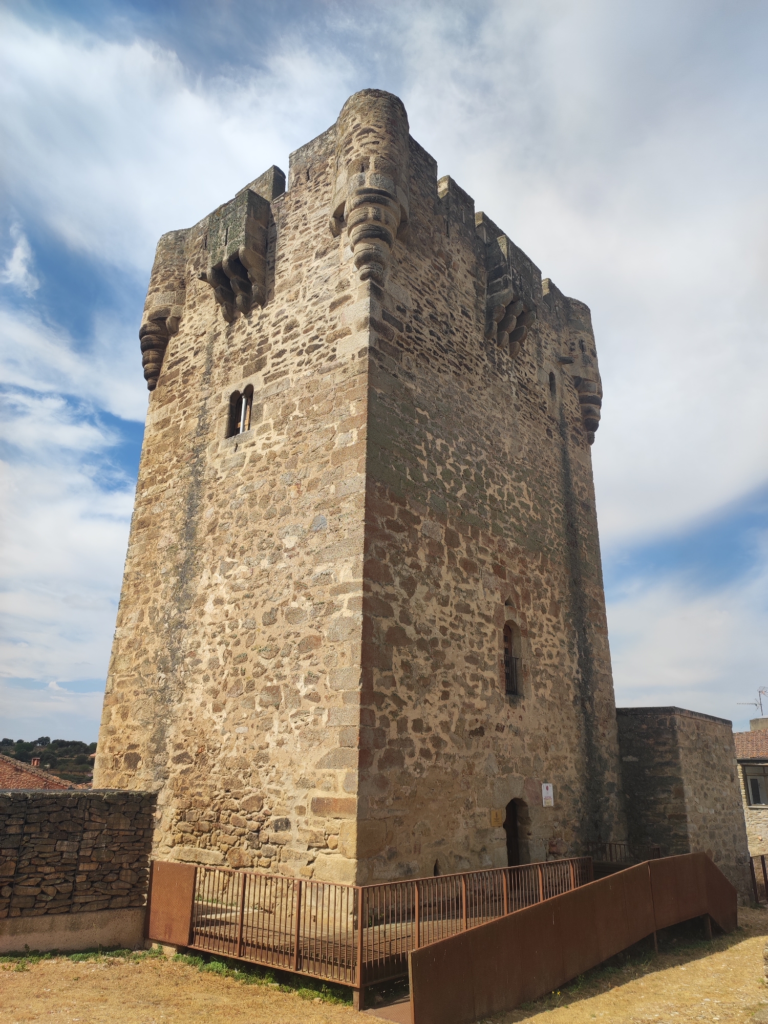 A medieval stone tower, a prominent feature of the town of Sobradillo, Spain, stands tall against a backdrop of a partly cloudy blue sky. The tower, constructed from rough-hewn stones of varying sizes and shades of brown and grey, has a square base and a crenellated top. Small, arched windows are visible at different levels on its facade. The lower section of the tower features an arched doorway. In the foreground, a rusted metal fence and a walkway lead towards the tower. To the left, a lower stone wall, also made of rough-hewn stones, separates the tower area from a dried, grassy field. In the distance, behind the tower, are glimpses of other buildings and a hilly landscape with some trees. The image captures a clear, daytime view of this historic structure.