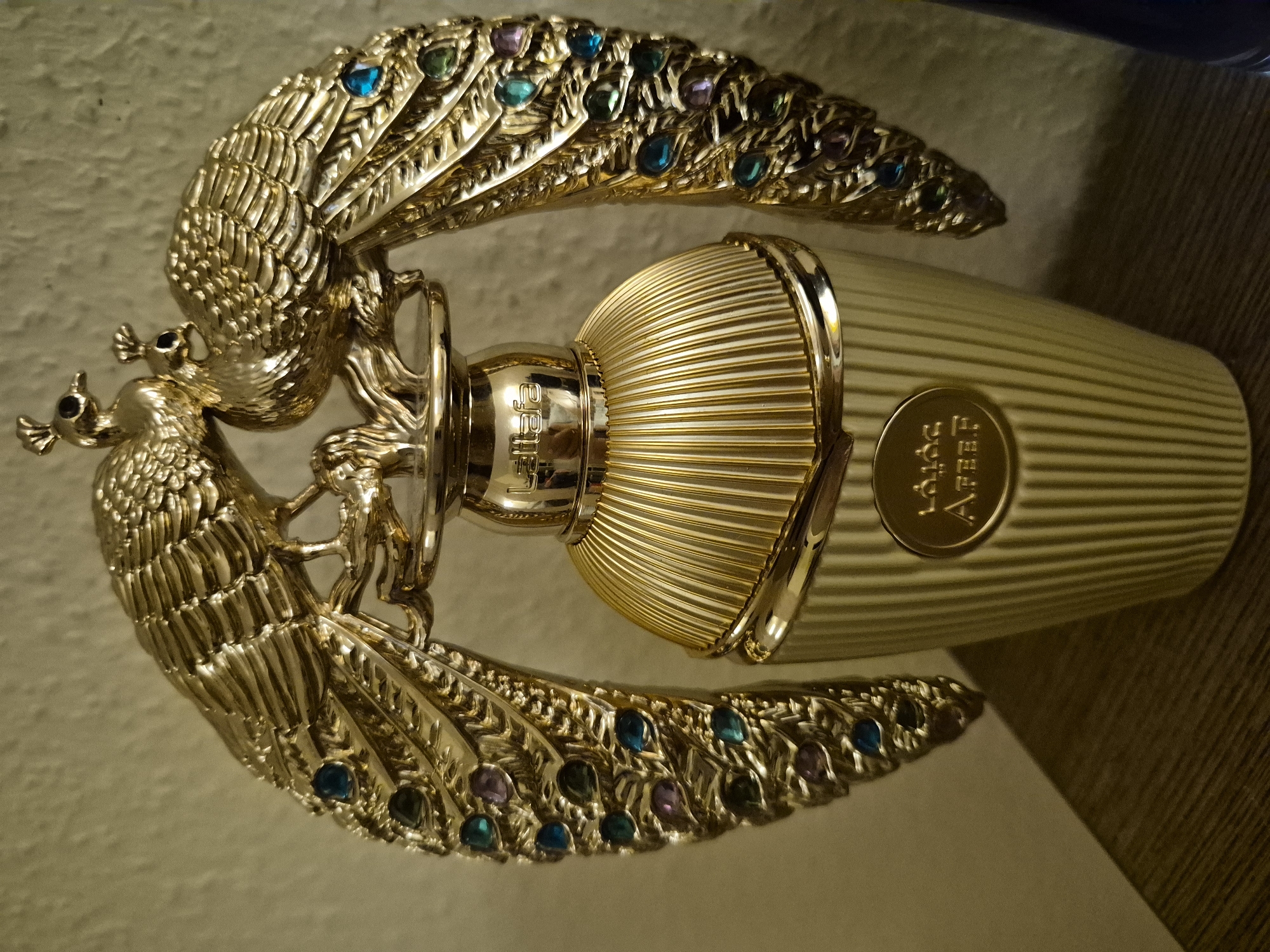 An ornate, gold-toned perfume bottle is displayed on a wooden surface against a textured wall. The bottle is topped with a decorative element featuring two gold peacocks with iridescent blue and purple accents on their wings. The bottle itself has a fluted design and a gold cap. A circular emblem with Arabic script and the word "AFEER" is affixed to the front of the bottle. The lighting is warm and casts shadows. The scene appears to be a product photograph, likely in a studio or display setting. There is no information in the image to indicate a specific city or location.