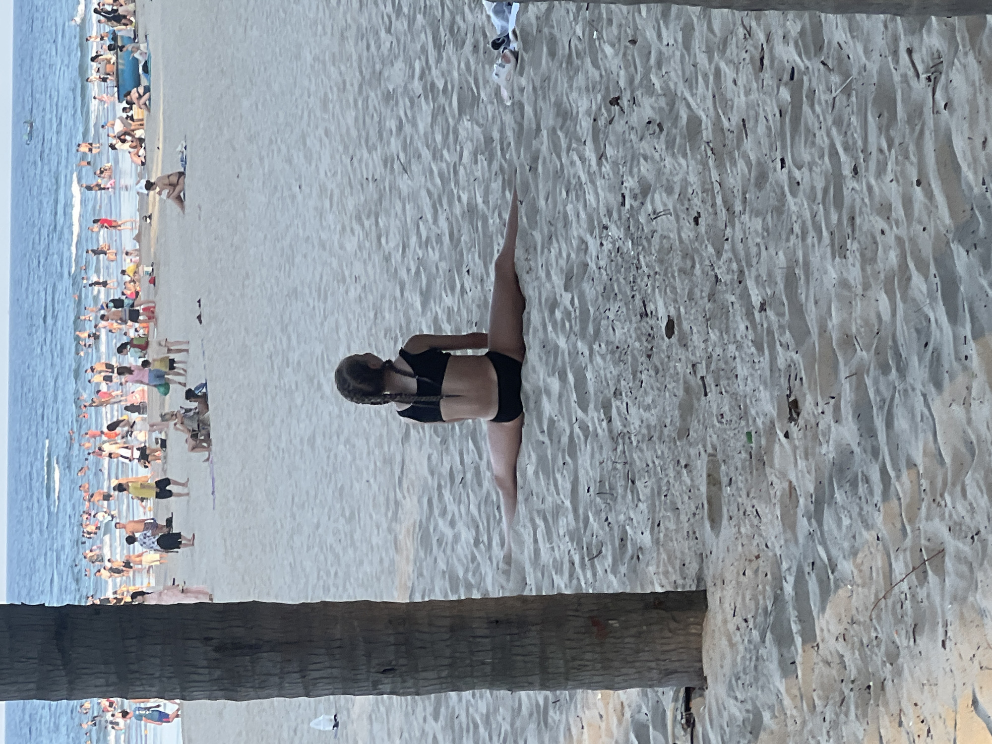 This media file shows a crowded beach in Da Nang, Vietnam. The image is oriented vertically, with the ocean and a large group of people on the left, and the sandy beach on the right. A palm tree trunk is prominently in the foreground, partially obscuring the view of the beach.
The water is blue and appears to have small waves, suggesting it is a calm day. People are scattered across the beach, some in the water, some walking along the shore, and many sitting or lying on the sand. The crowd is dense in some areas, indicating a popular spot. The lighting suggests it is daytime, possibly late afternoon given the warm tones.
In the middle of the frame, a young person in a black bikini is performing a leg split on the sand. Their hair is in two braids. They appear to be the central focus of the immediate foreground on the right side of the image.
Other details visible include various beach umbrellas and towels, as well as what appear to be some trash or debris scattered on the sand. The overall impression is a vibrant, busy beach scene typical of a popular tourist destination. No text is visible in the image.