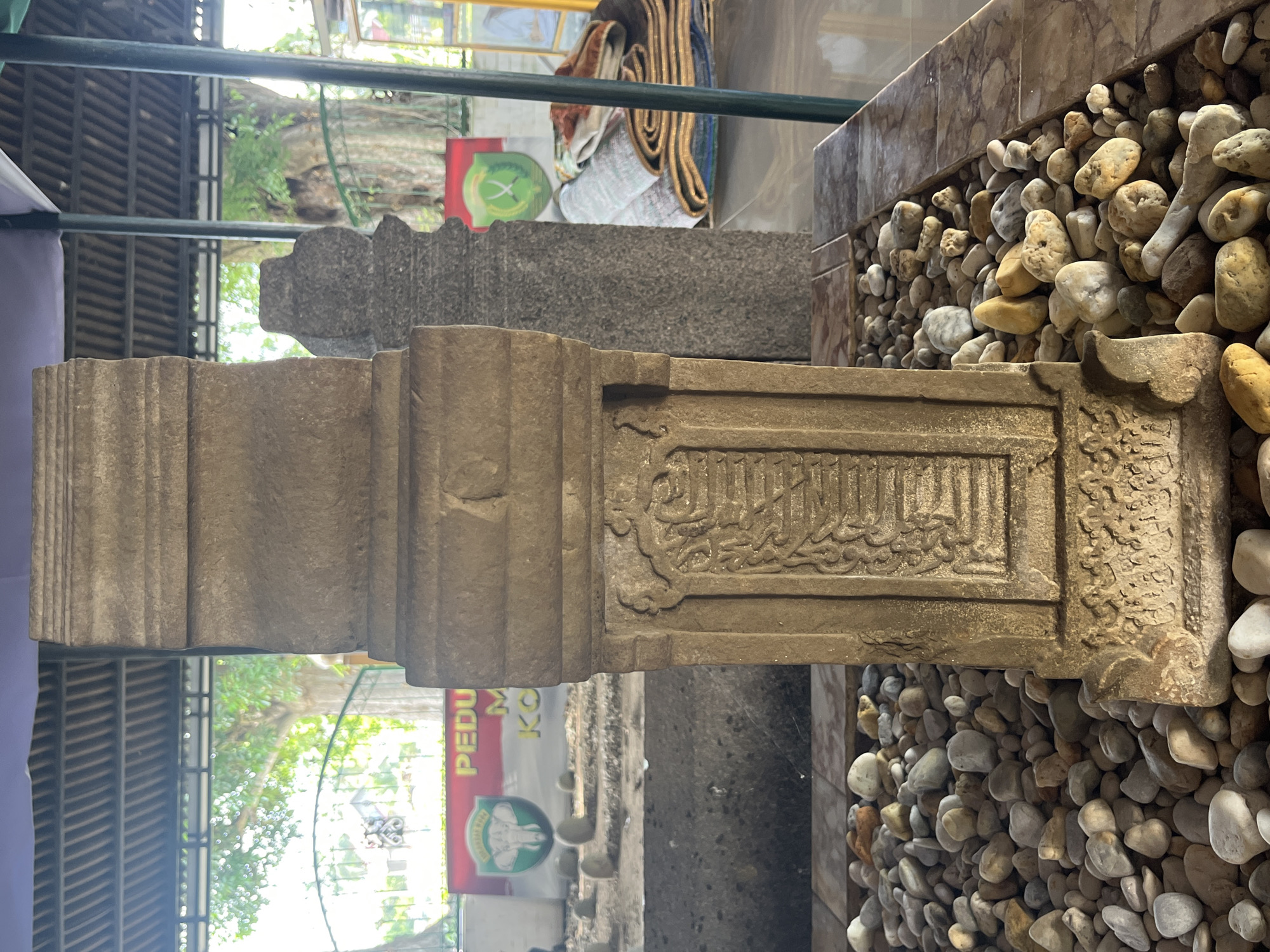 A stone monument, likely a tombstone, stands prominently in the foreground, adorned with intricate carvings. It is surrounded by a bed of smooth, rounded stones of various sizes and colors. The monument is situated outdoors, under a covered structure with a sloped, ribbed roof. In the background, other similar stone markers are visible, suggesting a historical burial site. Banners with logos and text are displayed behind the monuments. Lush green trees and foliage can be seen beyond the covered area. The overall environment appears to be a peaceful, historical site in Beuringen, Indonesia, possibly a cemetery or a sacred place of remembrance.