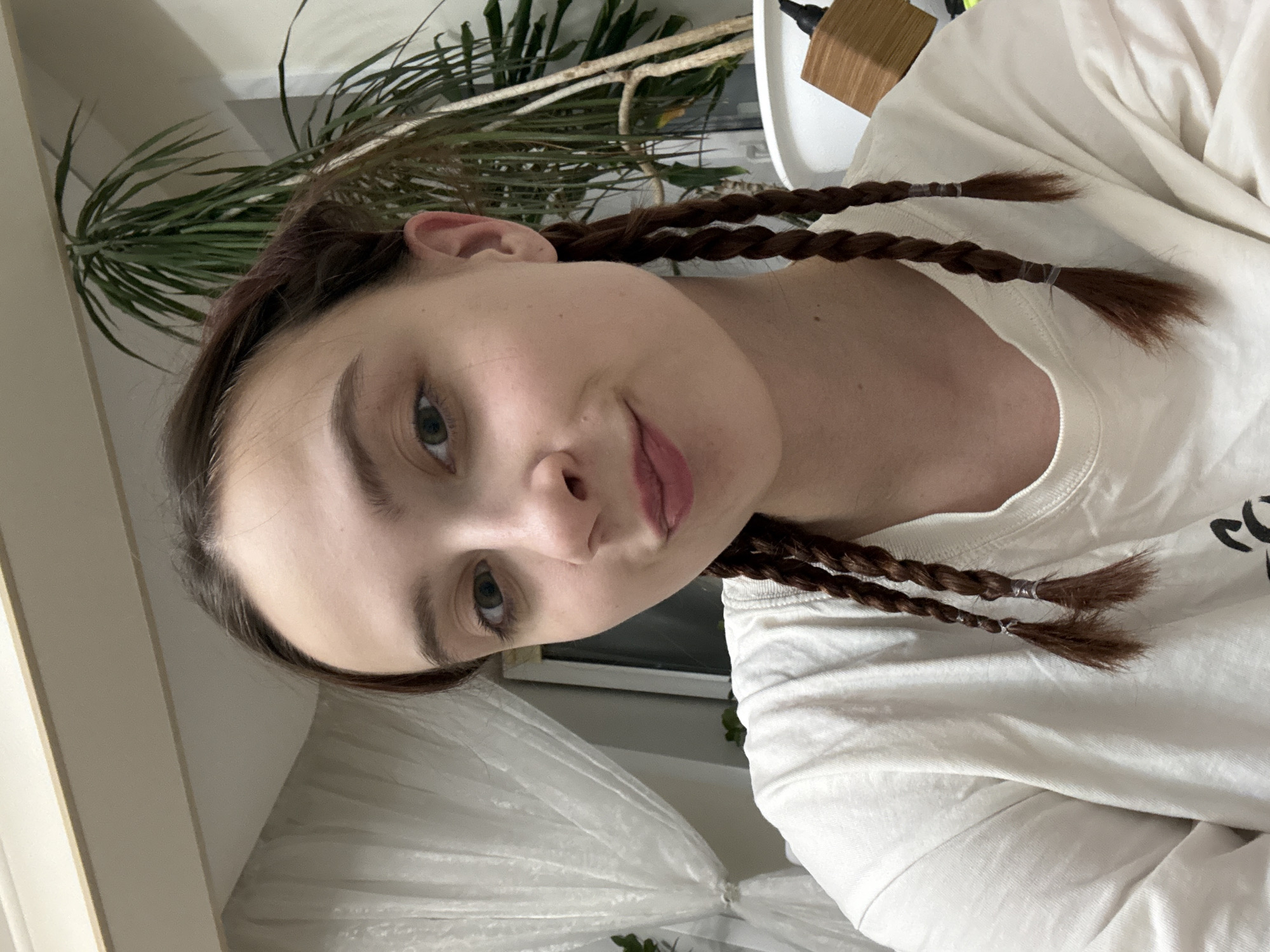 A young woman with brown hair braided into two plaits secured with clear elastic bands is looking at the camera. She is wearing a white t-shirt. The background shows a green plant, a window with white curtains, and a white wall. The location is not identifiable as the city of Vogulka, Russia based on the provided image.
