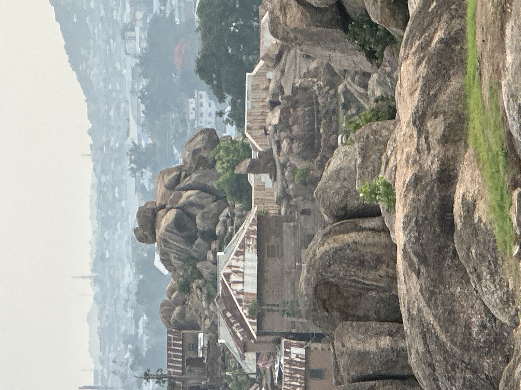 This image captures a rugged, rocky landscape, likely from an elevated perspective, showcasing a blend of natural rock formations and human habitation. Large, weathered granite boulders of varying sizes and rounded shapes dominate the foreground and midground, forming natural contours and small hills.
Nestled among these prominent rock formations are several small buildings, primarily houses or shelters. These structures feature corrugated metal roofs, some appearing oxidized with reddish-brown rust, while others are lighter in color. The walls of these buildings show a mix of materials, including what looks like brick or concrete, often built directly against or between the rock surfaces. Green foliage, including trees and bushes, is scattered throughout the scene, growing around and from the rocky crevices, adding pockets of vibrant color to the predominantly grey and brown palette.
In the background, a sprawling cityscape or town is visible, extending into the hazy distance. The distant urban area appears muted and somewhat indistinct due to atmospheric haze or dust, which also softens the lighting across the entire scene. The overall impression is of daytime, possibly during the morning or late afternoon, given the diffused light and lack of harsh shadows.
There are no people visible in the image, and no discernible text. The scene depicts a community living in close adaptation to a challenging, rocky natural environment, characteristic of parts of Jos, Nigeria.
