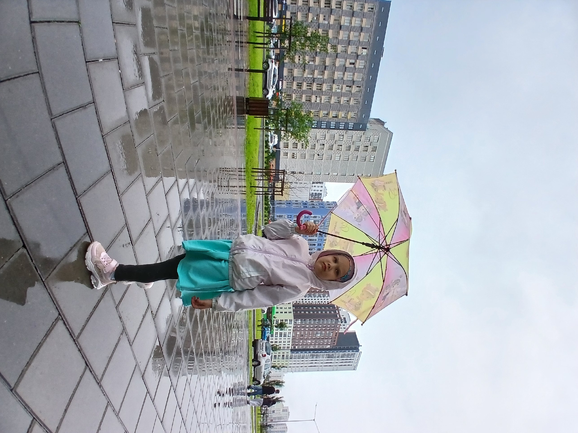 A young girl stands on a wet, paved sidewalk holding a colorful umbrella. She is wearing a light-colored hooded jacket, a teal skirt, black leggings, and pink sneakers. The umbrella is open and has a pattern of yellow, pink, and green sections, with cartoon-like figures visible on its surface. The girl is looking towards the camera with a neutral expression.
The scene appears to be outdoors, with modern, tall buildings in the background. The ground is wet and reflects the surroundings, suggesting it has recently rained or is raining. Several trees and some grassy areas are visible between the sidewalk and the buildings. Cars are parked or driving in the distance.
The overall atmosphere is somewhat somber due to the overcast sky and wet conditions, but the colorful umbrella adds a touch of brightness. The time of day is unclear due to the lighting, but it appears to be daytime. The location is indicated as Yekaterinburg, Russia. There is no visible text in the image.