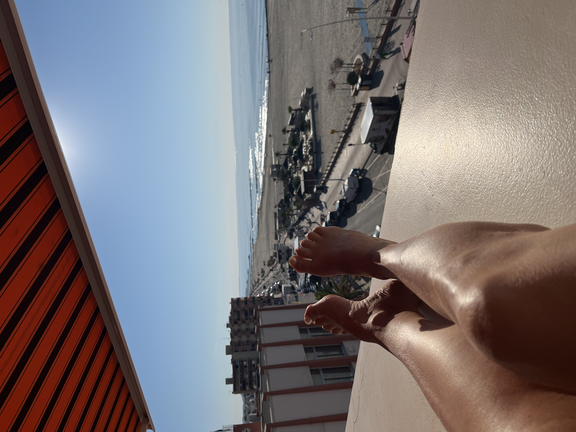 A first-person perspective shot shows a person's legs and feet relaxing on a balcony with a view of the Mediterranean Sea and the beach in Palavas-les-Flots, France. The awning above is orange with black stripes. The sea is calm and blue, with sunlight glinting off the surface. The beach is sandy with umbrellas and a few people walking. A road with cars and buildings runs parallel to the beach. The person's legs are tanned and bare.