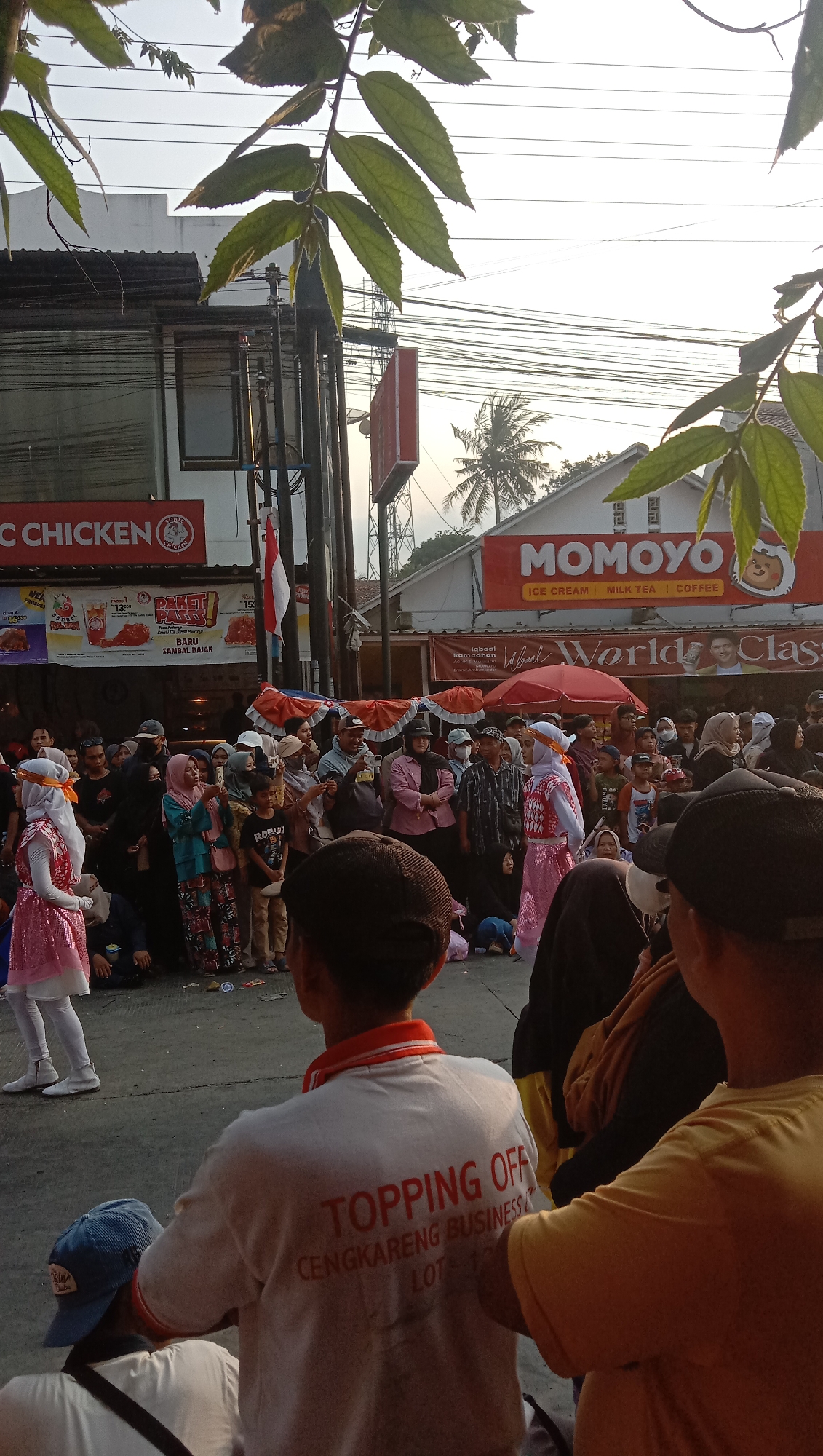 The image depicts a street scene in Jatisawit, Indonesia, likely during a public event or festival. A crowd of people is gathered, some observing a performance or procession. In the foreground, the back of a person wearing a white t-shirt with red lettering that reads "TOPPING OFF CENGKARENG BUSINESS LOT" is visible. To the left, a young person in a pink sequined outfit and white leggings stands. Behind them, several adults are watching, some holding umbrellas or flags. Storefronts with signage are visible in the background, including "MOMOYO" and "CHICKEN." Palm trees and electrical wires suggest an outdoor urban setting. The overall atmosphere appears to be festive and communal.