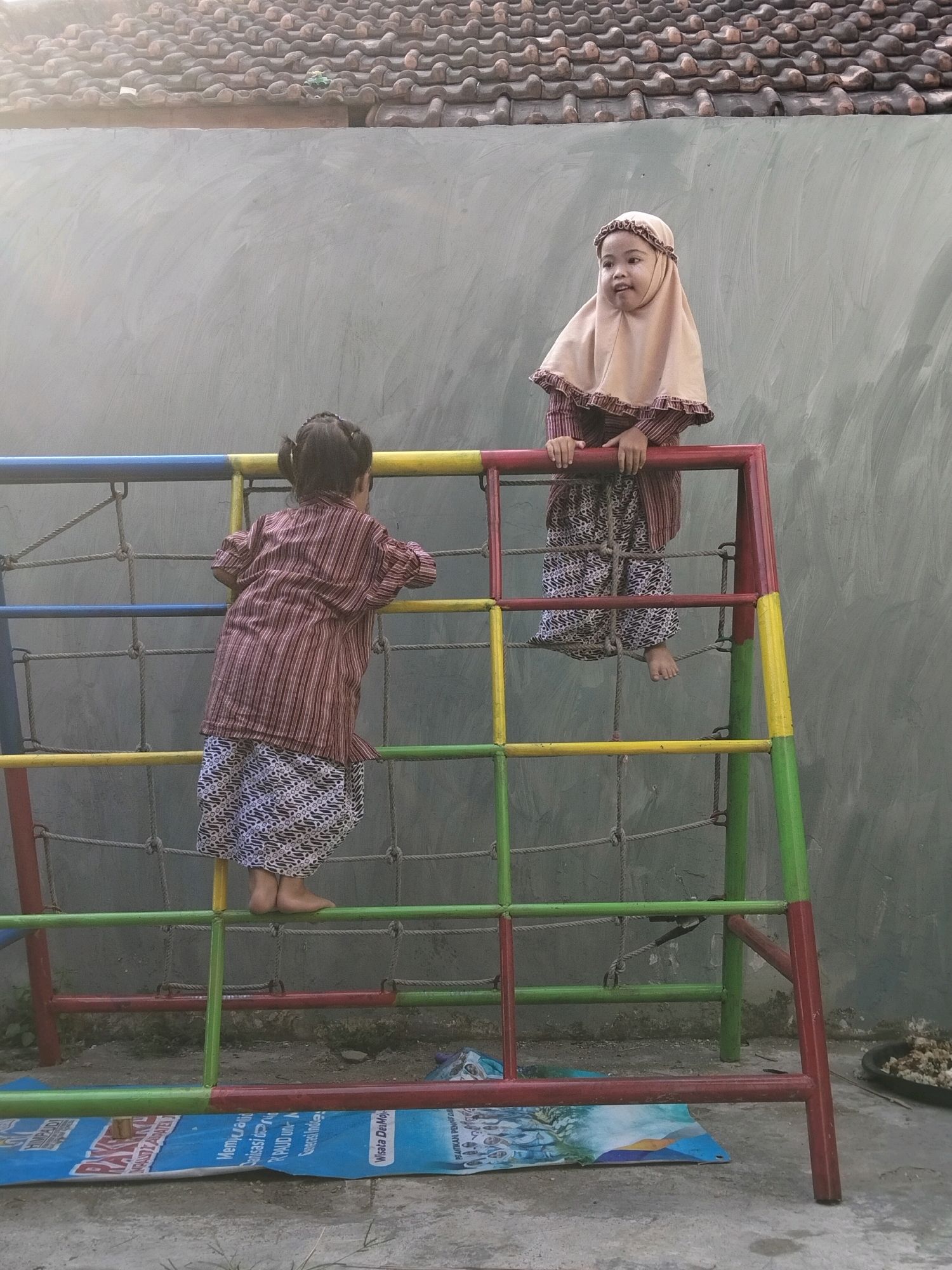 Two young girls are playing on a colorful climbing frame in front of a grey wall and a tiled roof. One girl, wearing a light brown hijab and a patterned dress, is standing on the top bar of the frame, looking to her left. The other girl, wearing a striped top and a patterned skirt, is climbing the frame from behind, facing away from the camera. The climbing frame has red, yellow, blue, and green bars and a net structure. A blue mat with text and images is on the ground beneath the frame. The environment appears to be an outdoor area, possibly a schoolyard or park, in Puskesmas Keboan, Indonesia.