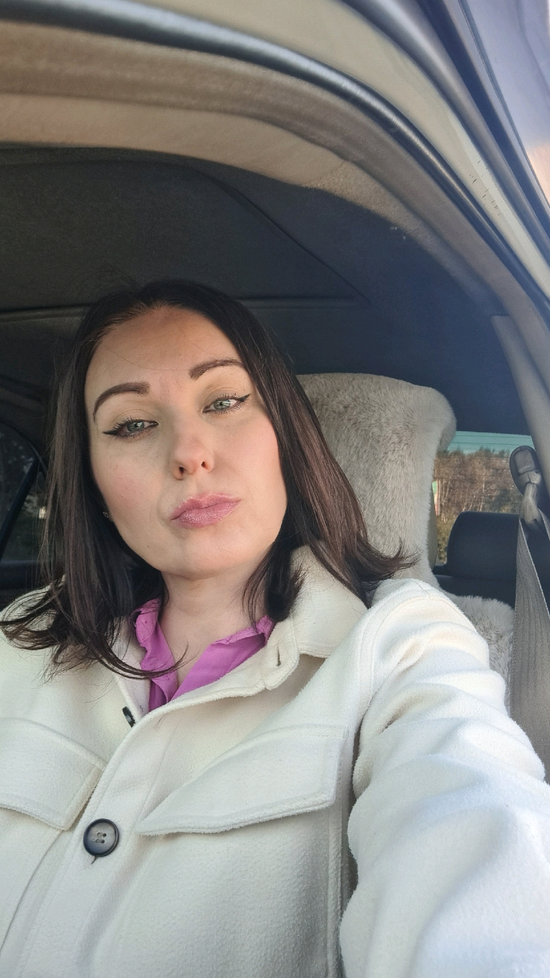 A dark-haired woman with light skin and green eyes is pictured inside a car. She is wearing a pink shirt and a cream-colored jacket. Her lips are pursed, and she is looking directly at the camera. The interior of the car is visible, including a fuzzy headrest in the background. The lighting suggests it is daytime. The image provides no specific details about the location within Irkutsk, Russia.