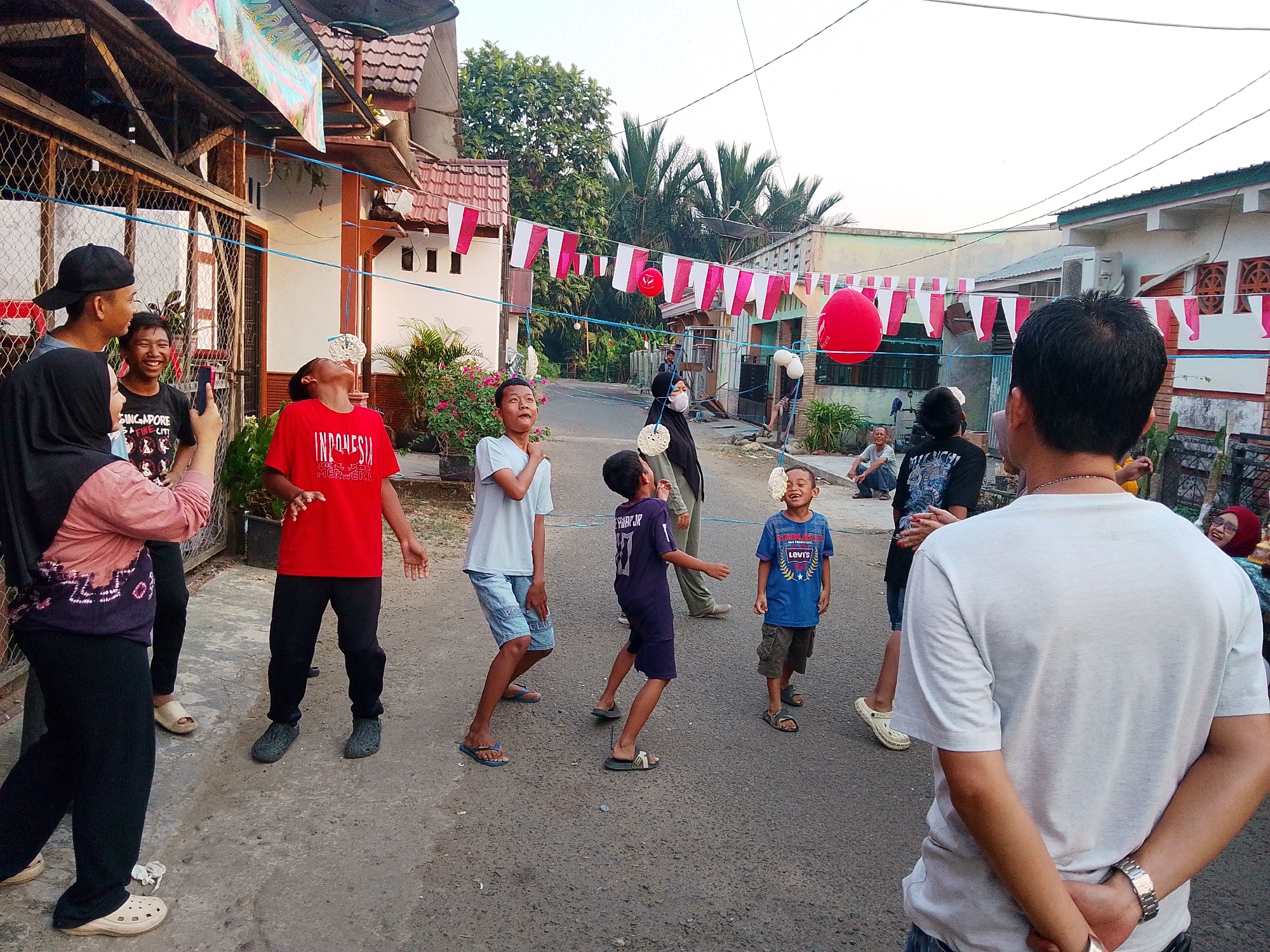 In a residential area of Barabai, Indonesia, a group of children and adults are gathered outdoors, likely for a community celebration. Red and white flags are strung across the street, suggesting a festive occasion, possibly Independence Day. Several children are engaged in a game, with one boy in a red shirt attempting to eat a cracker or snack suspended from a string. Another boy in a blue t-shirt watches with a wide smile, also with a suspended snack near his face. The street is paved, and houses with tiled roofs line both sides. A few adults are present, observing the activities, with one person in the foreground facing away from the camera, wearing a white t-shirt. The lighting suggests it is late afternoon or early evening.
