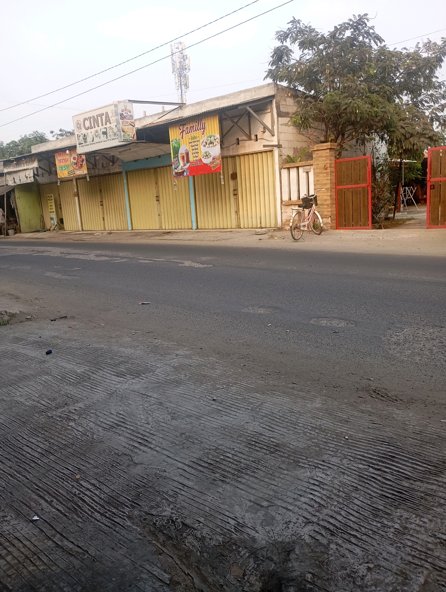 The image depicts a street scene in Babelan, Indonesia, characterized by a row of closed shops with yellow roll-up doors. A white sign with red lettering, reading "CINTA," is mounted above one of the shops, accompanied by another sign with a picture of a motorcycle. To the right, a colorful banner advertising "Family" food products hangs from another shop. A pink bicycle with a basket is parked on the sidewalk in front of a brick pillar and a red wooden gate. The street in the foreground is asphalt with visible cracks and textured concrete. In the background, a cellular tower is visible against a hazy sky. The scene appears quiet and possibly during off-hours due to the closed businesses.