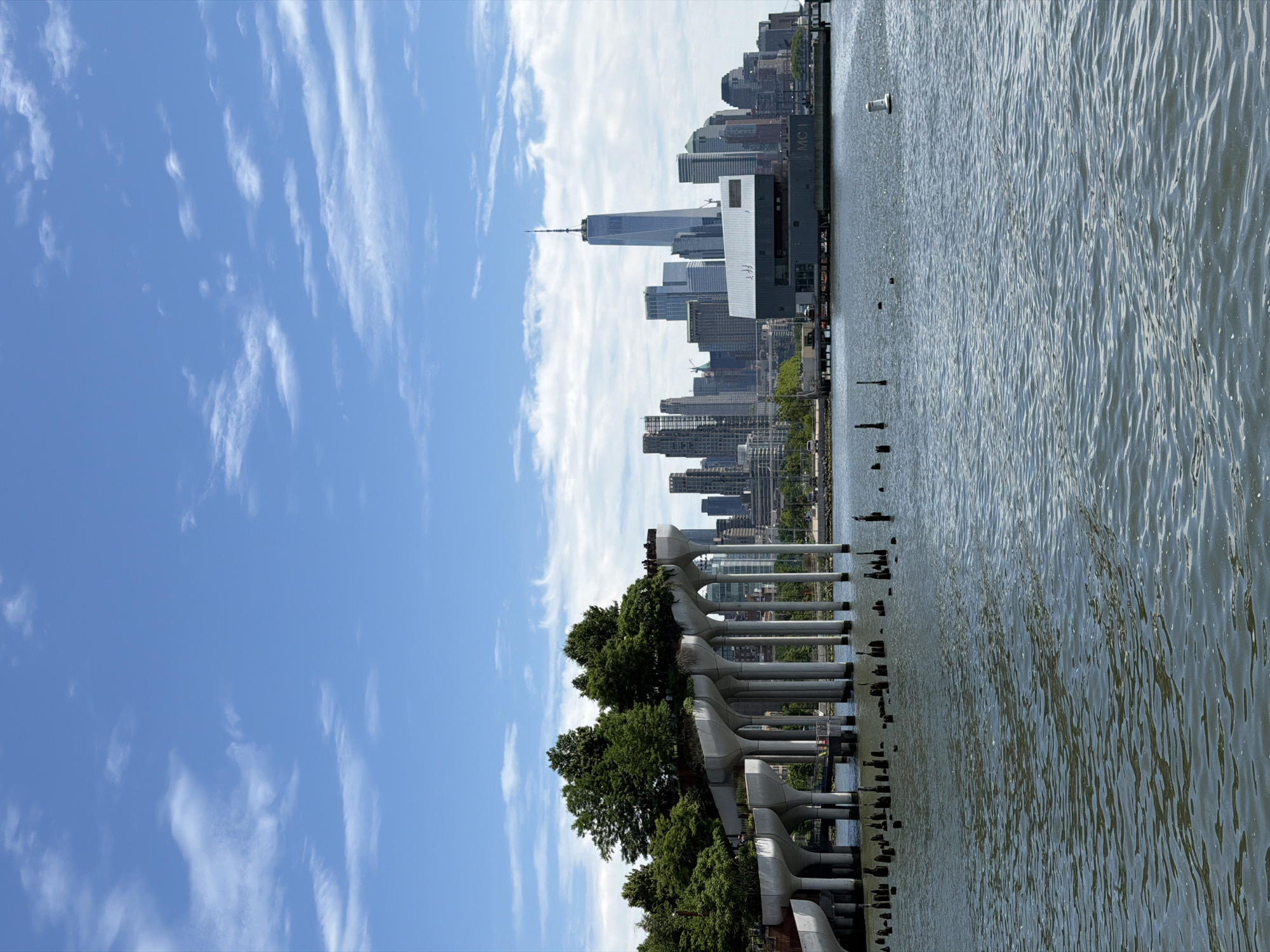 The image is a landscape photo taken outdoors. The viewpoint is from the ground level, with the camera tilted 90 degrees to the left. The sky occupies approximately half of the image, displaying a bright blue color with sporadic, thin, white clouds scattered across it.
In the foreground, there is a section of a pier or walkway structure composed of gray concrete. The structure is partially obscured by lush green trees and foliage. Wooden pilings, likely remnants of an older structure, are visible near the water's edge.
The other half of the image features a body of water, presumably a river or bay. The water appears slightly choppy with visible waves.
In the background, the skyline of New York City is apparent. The most prominent building is the One World Trade Center, its distinctive antenna reaching into the sky. Other skyscrapers and buildings populate the skyline, typical of the urban landscape of Manhattan.
There are no people visible in the image. The setting suggests a waterfront park or recreational area with a view of the Manhattan skyline.