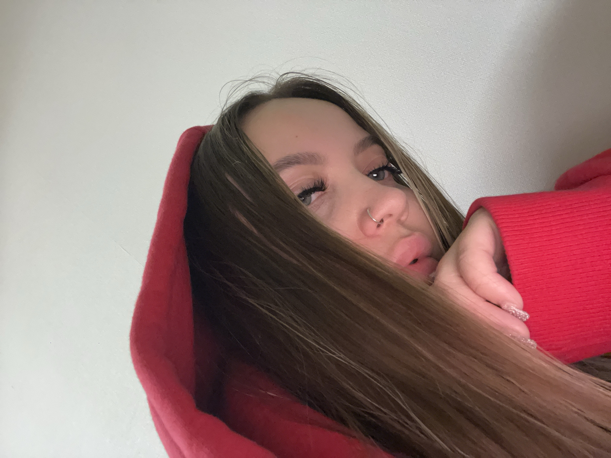 The image is a close-up, angled portrait of a young woman, likely a selfie. She has long, straight, medium brown hair that partially covers her face, framing her left eye and nose. She is wearing a bright red hoodie, with the hood pulled up around her head.
Her visible left eye appears to be blue or grey, accentuated by prominent dark eyelashes. A silver hoop piercing is visible on her right nostril. Her lips are full and slightly parted. One of her hands, adorned with long, light-colored, possibly glittery fingernails, is positioned near her chin, partly obscured by her hair and the red hoodie's sleeve.
The woman is looking directly towards the camera with a neutral to slightly pensive expression. The background is a plain, light-colored wall, possibly white or off-white, with a subtle textured surface. The lighting is soft and even, suggesting an indoor setting. No specific time of day or weather conditions are discernible from the image. The provided location context is Krasnodar, Russia, though this is not visually evident within the photograph. There is no visible text.