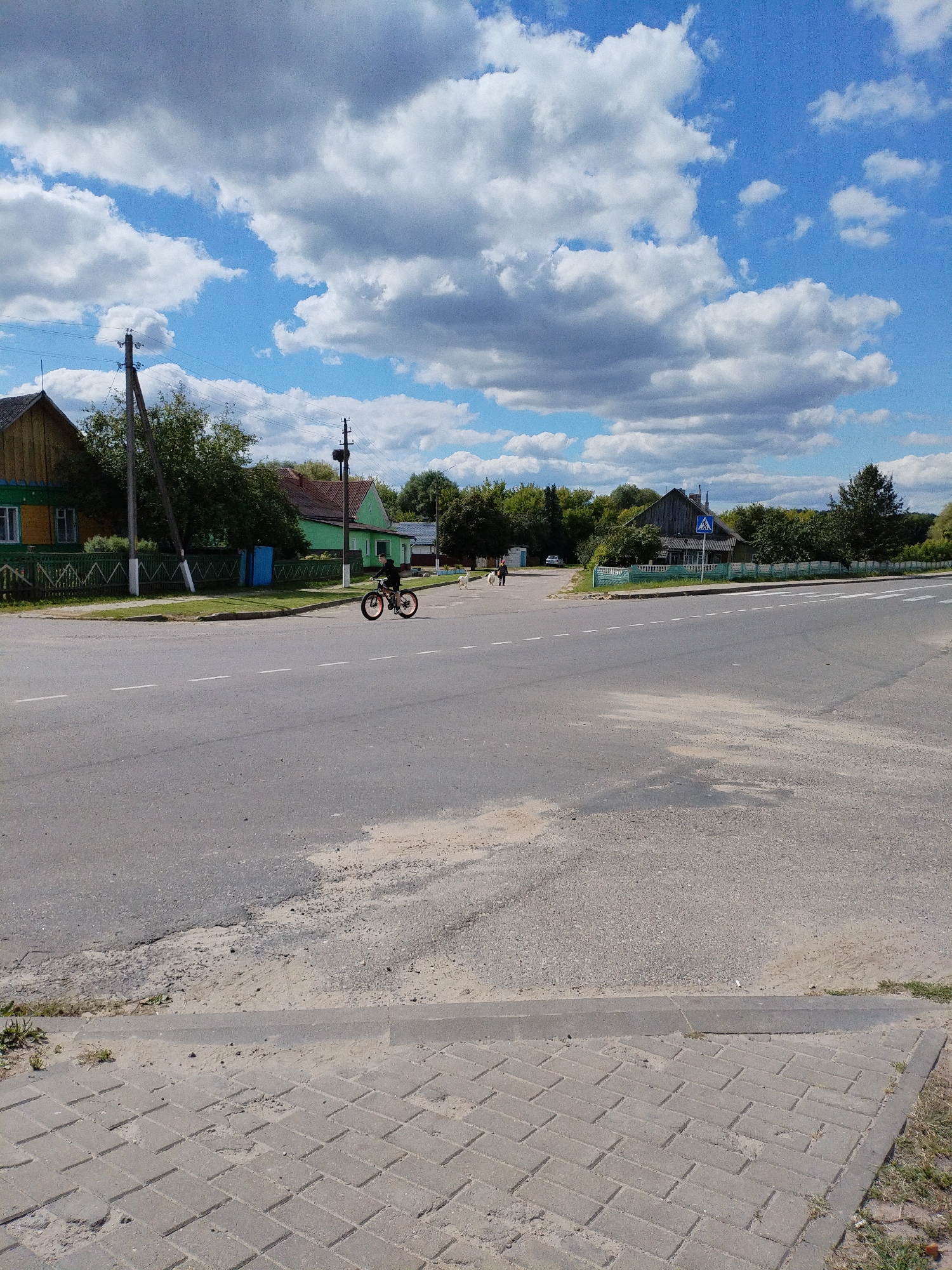 An eye-level, full outdoor shot captures a road intersection in Bialynichy, Belarus, under a bright, partly cloudy sky. The road, made of asphalt, has two white dashed lines marking lanes. To the left, a paved sidewalk transitions into a brick pathway near the curb. Beyond the sidewalk, a green wooden fence borders a yard with a yellow and green house. Further down, a long, low green building is visible. A single person is riding a black bicycle with orange rims on the road. In the distance, two people and a white dog are walking near a pedestrian crossing sign. Trees and additional houses line the street in the background.