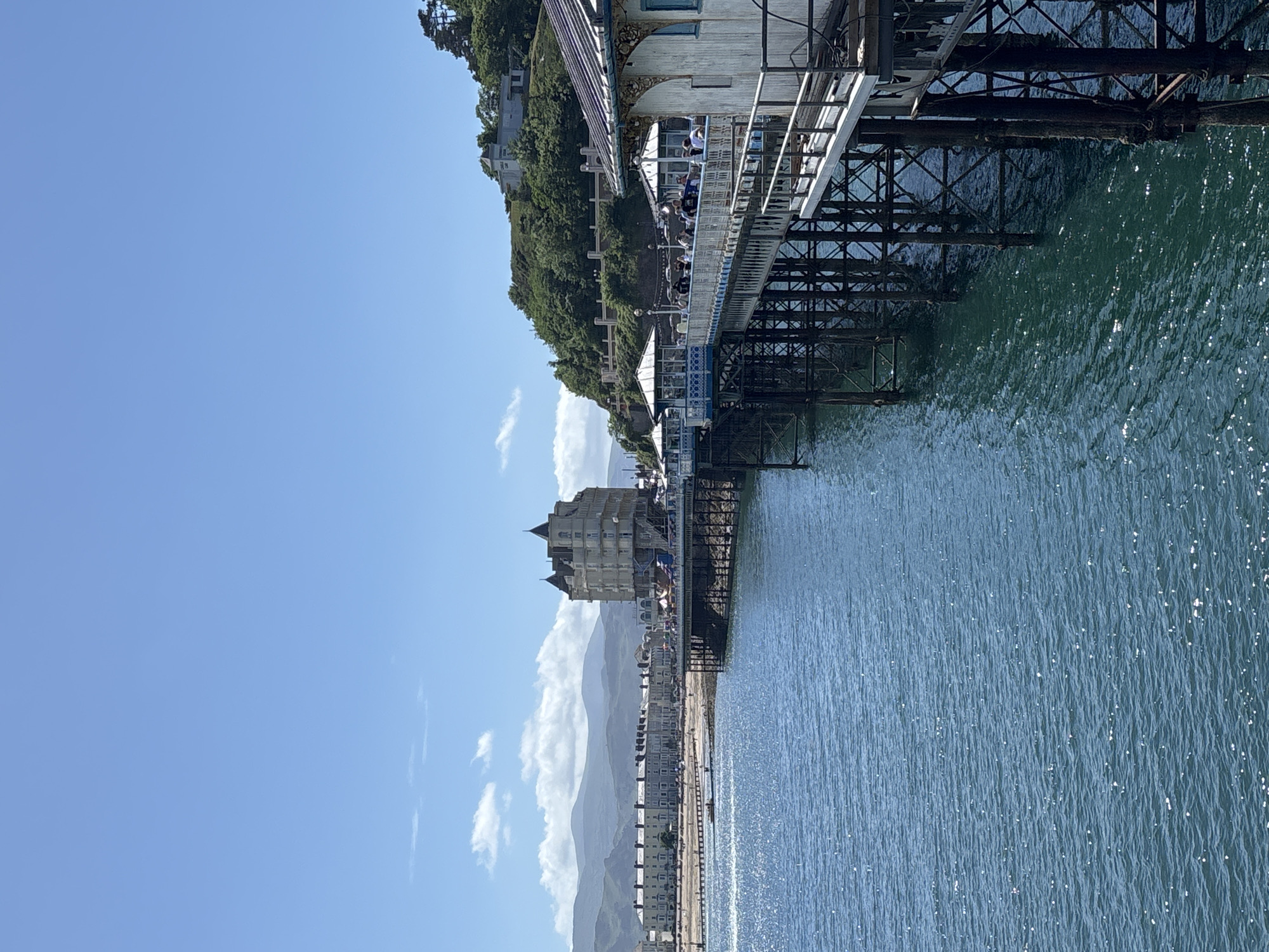 The image captures a sunny day in Llandudno, United Kingdom. A pier stretches into the sea, supported by metal framework. People can be seen walking on the pier. The shoreline features buildings and a distant mountain range under a clear blue sky with scattered clouds. The sea is shimmering with sunlight.