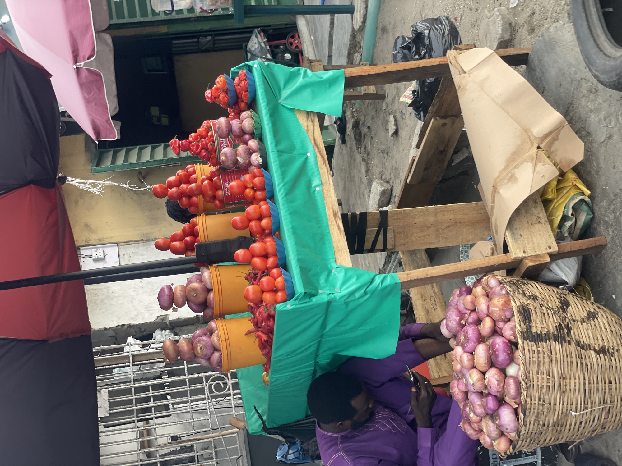 This image captures an outdoor market scene, likely a street stall, in Ado, Nigeria. The primary focus is a display of fresh produce and a man seated beside it.
The stall consists of a wooden structure, partially covered with a bright green tarpaulin. Arranged vertically on this structure are several yellow buckets, each overflowing with different vegetables. Red tomatoes are visible in multiple buckets, while others hold red peppers (likely Habanero or similar) and purple onions.
A man, dressed in a purple long-sleeved garment, sits on the ground to the right of the stall, leaning against its structure. He is engrossed with a mobile device held in his hands. Beside him, a large woven basket overflows with numerous purple onions, some spilling onto the ground.
In the background, a building with a dark facade and windows is partially visible, along with a metal security grille. An electrical outlet is fixed on a light-colored wall. Two umbrellas, one pink and one black and red, are positioned to provide shade for the stall. The ground is a mix of rough concrete and dirt, with black trash bags and a discarded tire indicating a bustling urban environment.
Visible text includes "SONIA" and "ONI" on product labels near the tomatoes and peppers, and "SURFACE PREPARATION" faintly seen on a bag near the basket of onions. The scene depicts a typical daytime market activity, vibrant with colors from the produce and stall coverings.