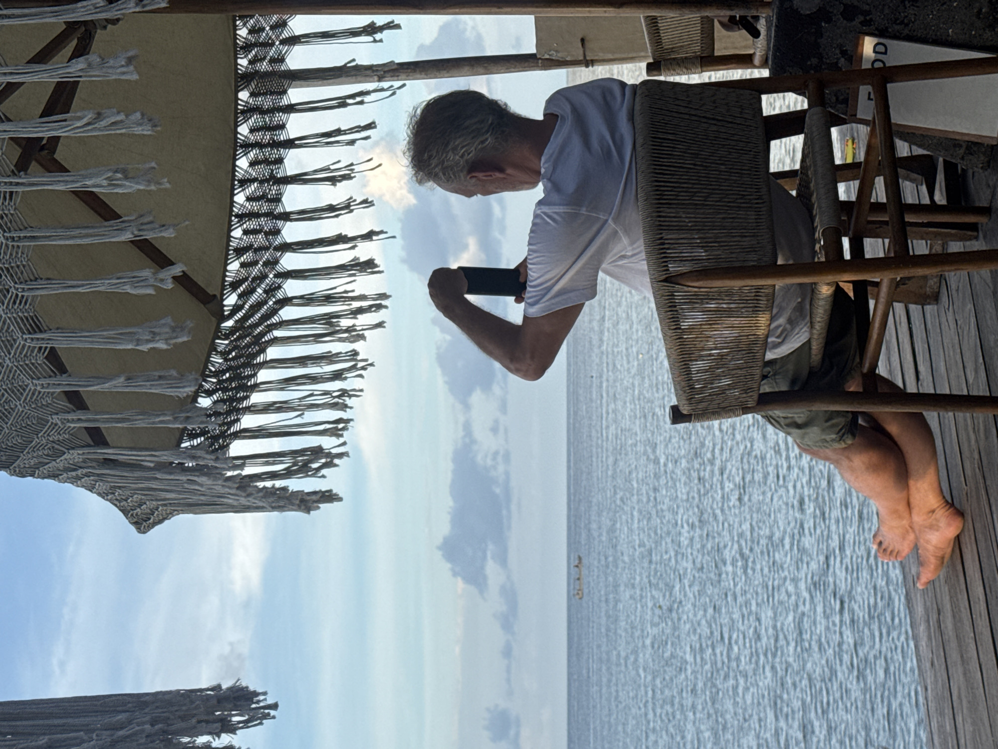 A man with short gray hair sits in a woven chair on a wooden deck and takes a photograph of the ocean with his phone. The man wears a white t-shirt and olive-colored shorts. His bare feet rest on the wooden deck, which appears to be part of a stilt structure over the water. The ocean is in the background with a faint boat in the distance. Above the man and to the left is a fringed sunshade with a geometric pattern. The sky is visible through the fringe, filled with white clouds. Based on the information provided, this scene is likely taking place in Nusa Penida, Indonesia.