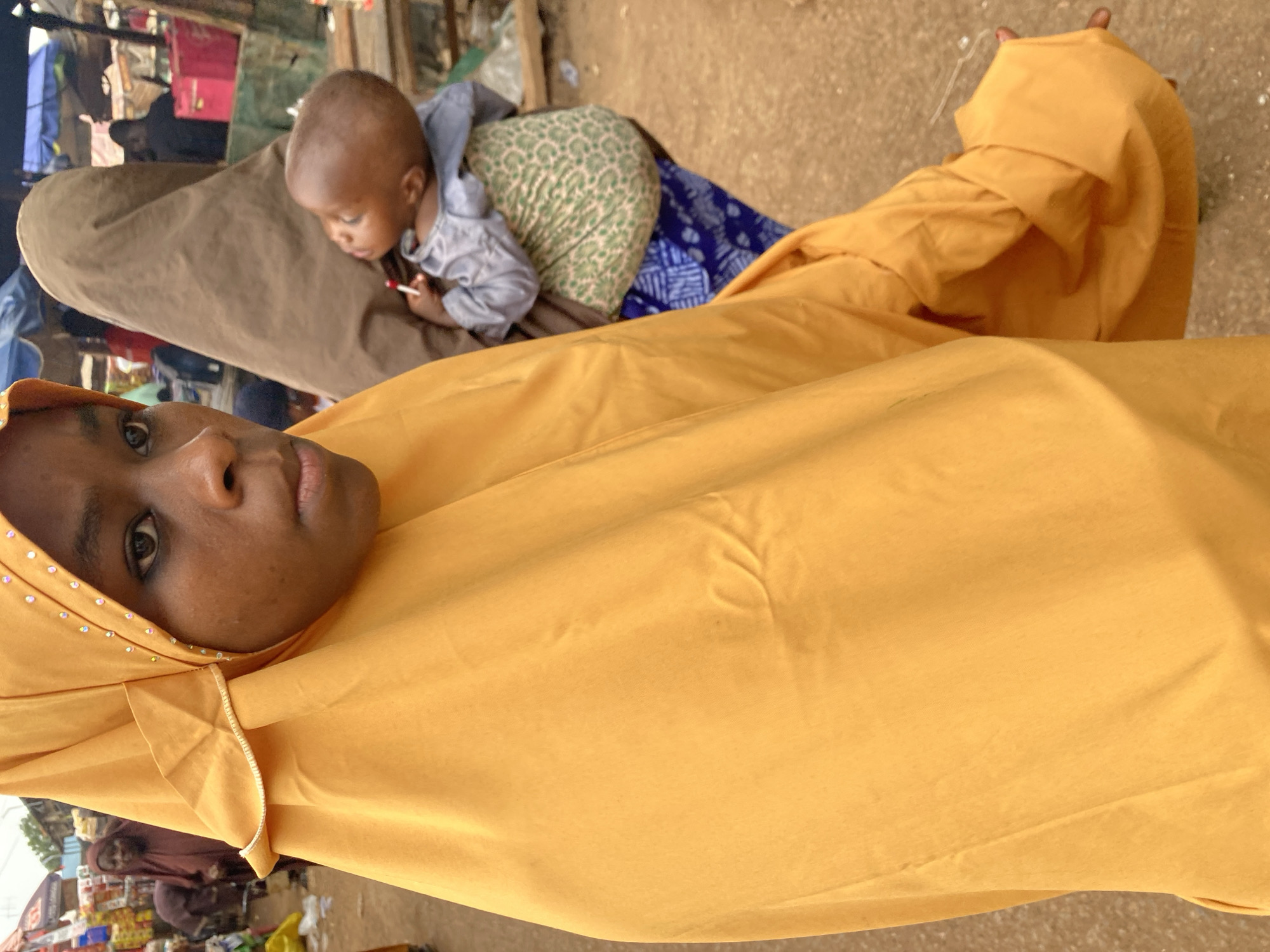 In a market in Bukuru, Nigeria, a young woman wearing a bright orange hijab, adorned with small rhinestones along the edge, stands in the foreground. Her gaze is directed slightly off-camera to the right, with a neutral expression. Behind her, another woman, mostly obscured by her own brown hijab, carries a baby on her back. The baby, facing left, is wearing a light blue garment and has a lollipop in its hand. The baby is wrapped in a green and black patterned fabric. The background reveals a bustling market scene with stalls, merchandise, and other people, suggesting a lively commercial environment. The ground is dirt or gravel.