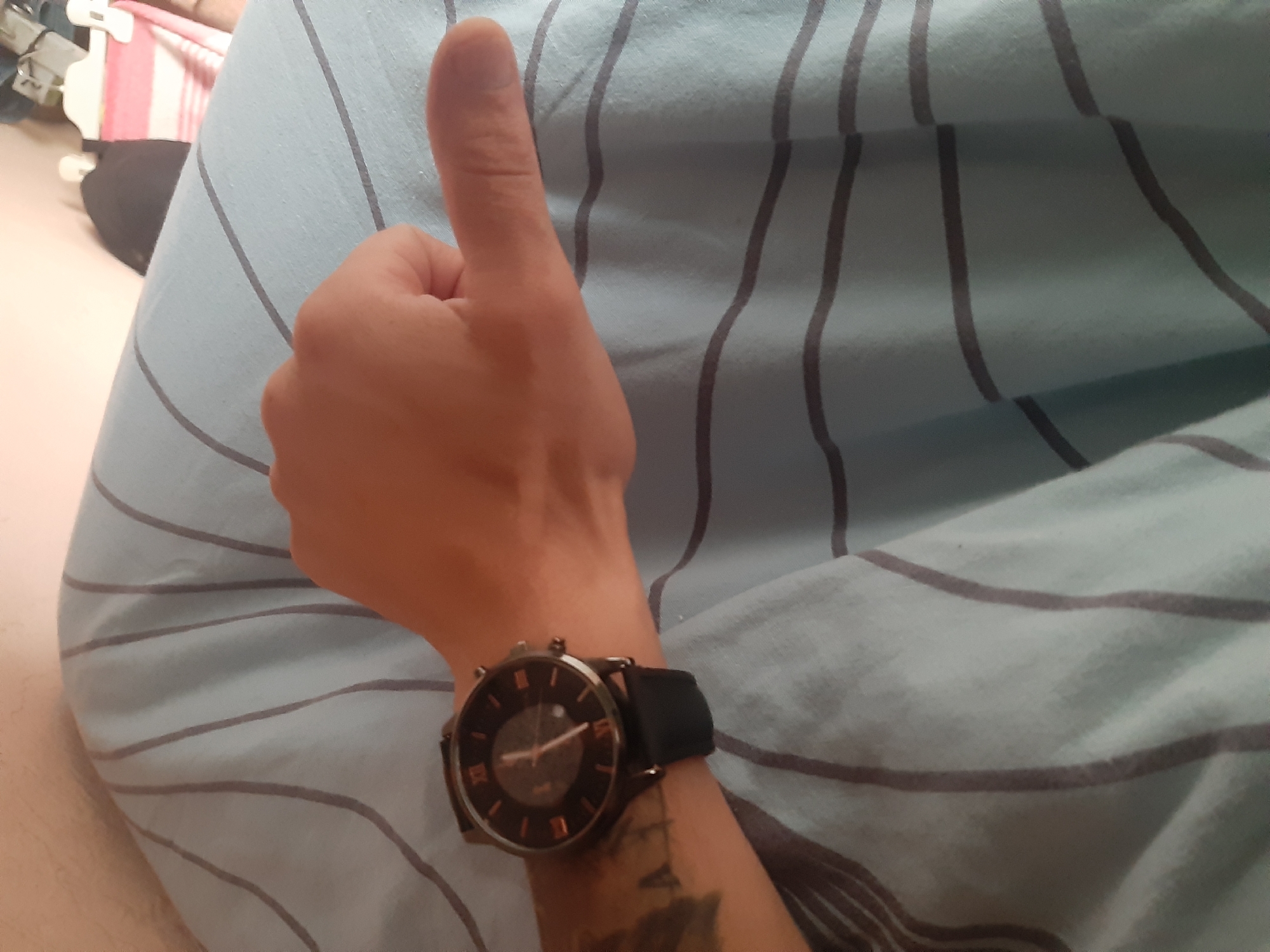 A person's left arm, wearing a black watch with a dark face and rose gold roman numerals, rests on a light blue striped blanket. The thumb of the hand is extended upwards in a thumbs-up gesture. The background is blurred, but appears to be indoors, possibly in Cricova, Moldova, given the context.