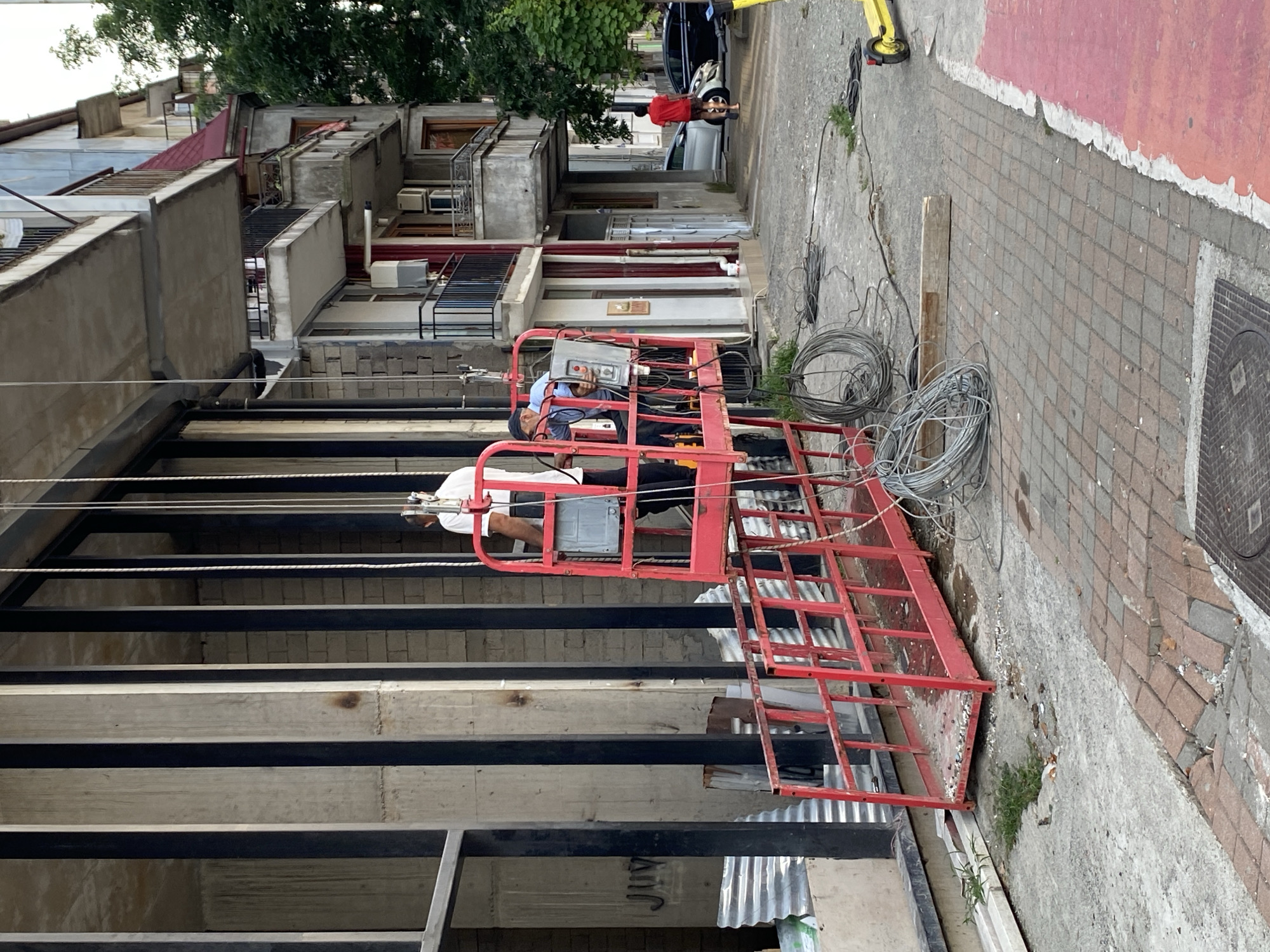 The image depicts an outdoor construction or maintenance scene in Batumi, Georgia. Two men are visible on a red suspended platform lift, which is raised against a building's facade. The building features a grid of large, light-colored vertical concrete pillars interspersed with darker vertical elements. The platform lift is equipped with control panels and what appear to be tools or equipment. Below the lift, coiled wires and construction debris are scattered on the ground. The ground surface transitions from concrete to a patterned brick walkway, with a section painted red. In the background, balconies with railings are visible on an adjacent building, and trees line the street. Two cars and a yellow scooter are parked further down the street. The overall impression is of ongoing work in an urban environment.