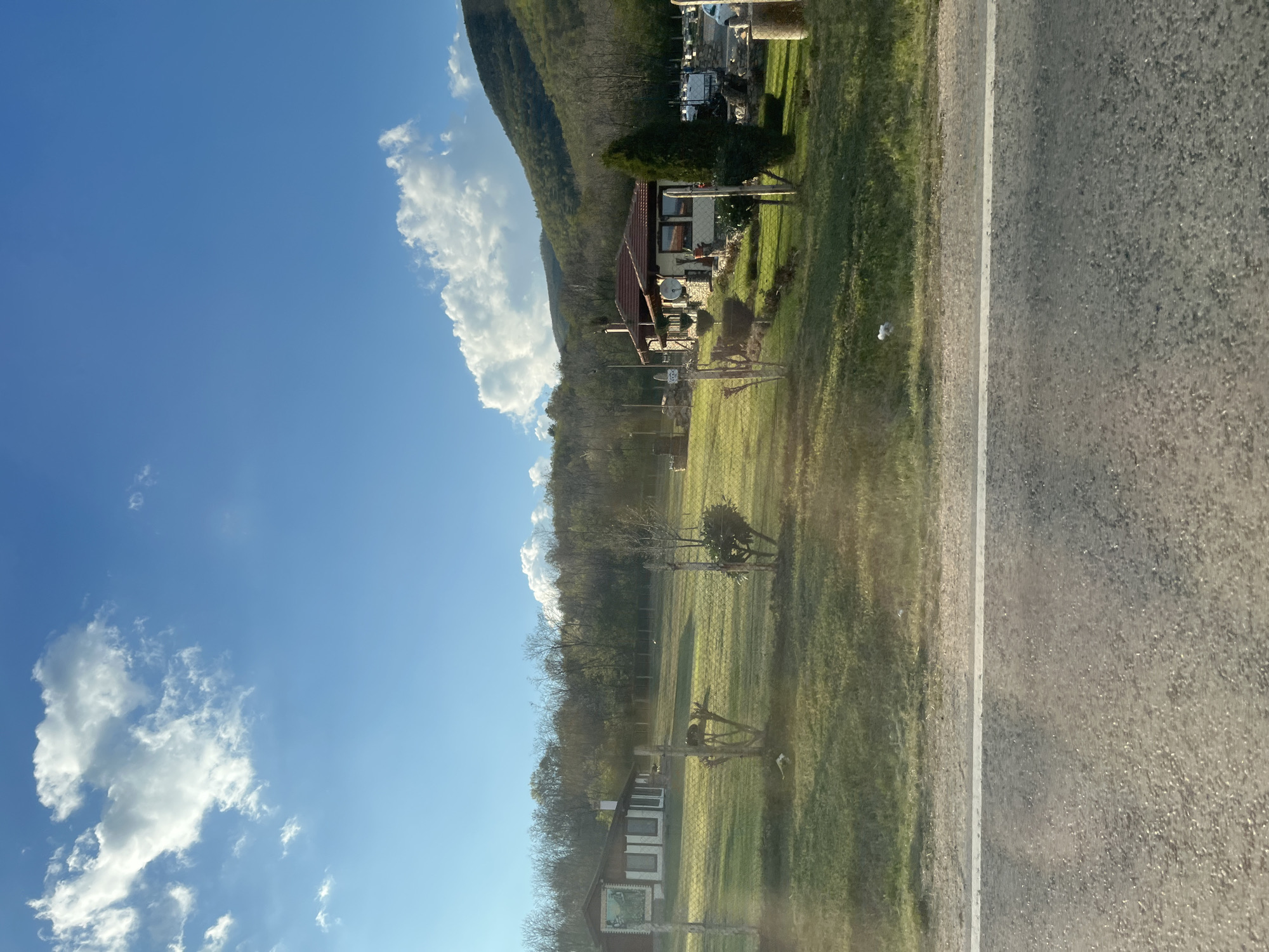 The image depicts a rural scene likely taken from a moving vehicle on a road in or near Osetenovo, Bulgaria. To the right is a two-lane road with one visible white line marking separation. Beyond a grassy verge, a chain-link fence encloses a large green field with scattered trees and wooden structures. A single-story house with a red-tiled roof, satellite dish, and garden is visible further up the slope, nestled against a tree-covered hillside. The sky above is clear blue with scattered white clouds. The scene is generally peaceful and indicates a landscape with residential and natural elements, consistent with rural areas.