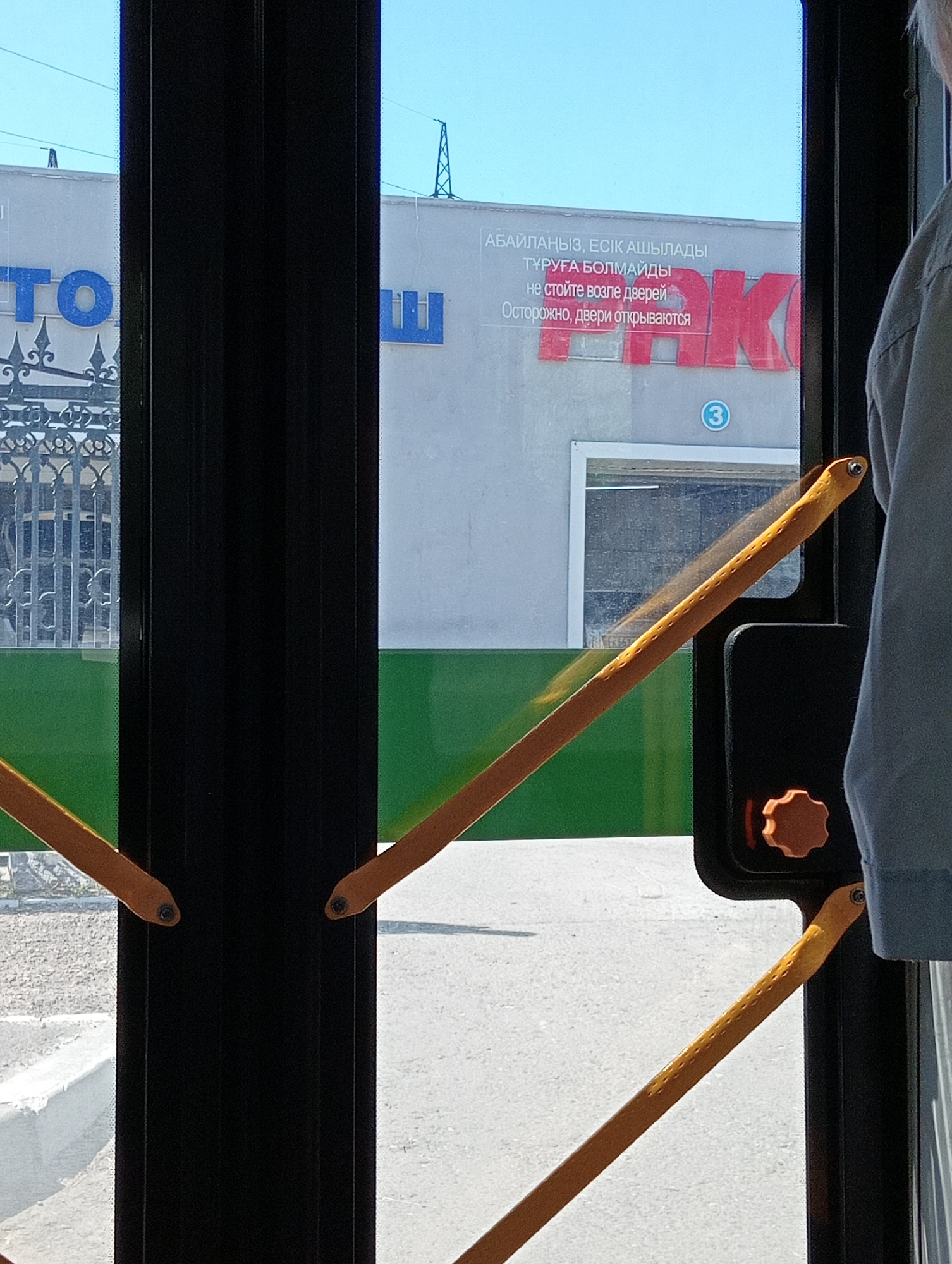 The image shows a view from inside a bus or tram, looking out through the doors. In the foreground, there are two yellow handrails, one extending diagonally from the bottom left to the middle right, and another from the bottom right to the middle left. These handrails have small indentations along their length. A black vertical pillar is visible between the two doors. To the right, a person's grey sleeve is visible.
Through the glass doors, a building with a light-colored facade is visible. Large red letters forming the word "РАК" are affixed to the building. Above these letters, there is text in what appears to be Cyrillic or a similar script, with the phrase "не стойте возле дверей" and "Осторожно, двери открываются" clearly legible, indicating "do not stand near the doors" and "careful, the doors are opening." A blue circular sign with the number "3" is also visible on the building. Further in the background, there are glimpses of a decorative metal fence and a transmission tower. The sky is clear and blue. The environment appears to be urban.