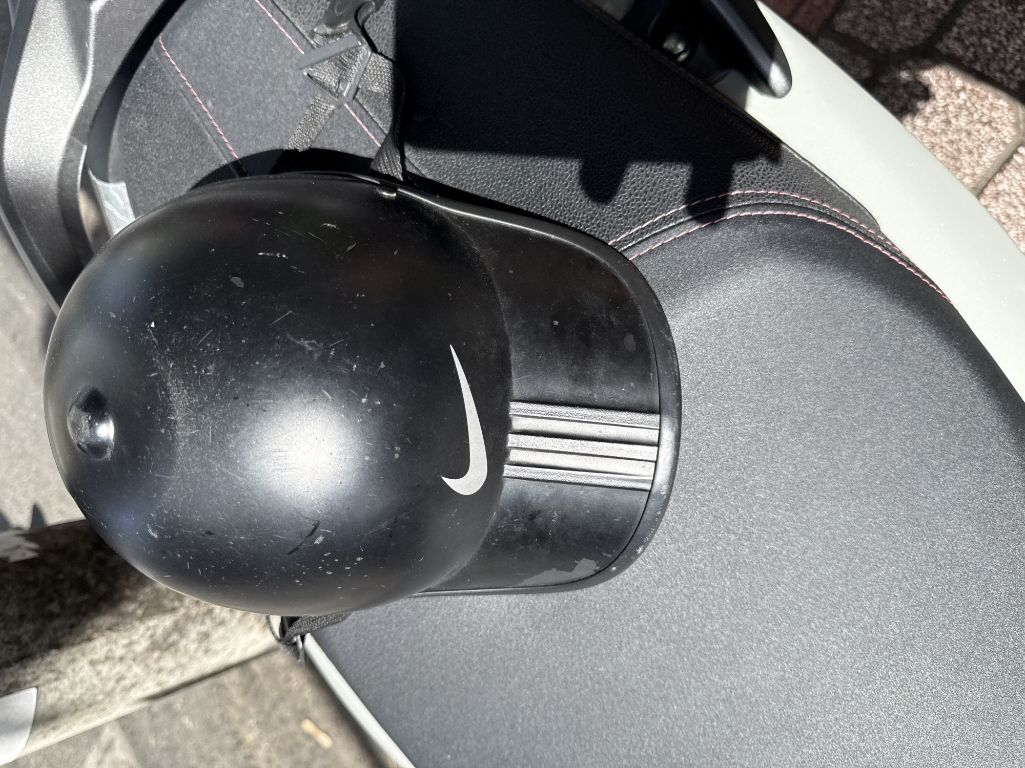 The image shows a black Nike baseball cap helmet placed on the seat of a white scooter. The helmet is positioned on the left side of the scooter seat, with its brim facing towards the bottom of the image. The Nike logo is visible on the front of the helmet. The scooter seat is black and textured, with visible pink stitching along the edges. The scooter is parked outdoors, with a blurred background that appears to be a sidewalk and a pillar. The lighting suggests it is daytime, with sunlight casting shadows. The scooter is on a cobbled surface, which is consistent with some areas in the city of Nha Trang, Vietnam, particularly in tourist-frequented areas or older sections of the city. There are no people visible in the image.