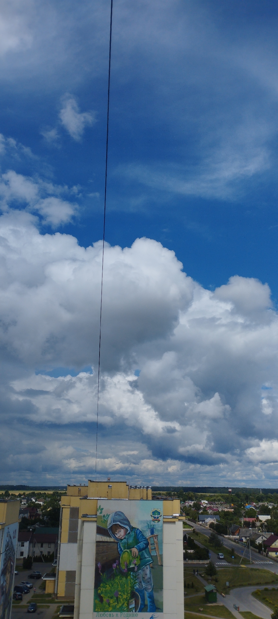 This image captures a vertical view looking upwards towards a bright blue sky filled with dramatic, puffy white and grey clouds. A thin, dark line, likely a cable or wire, bisects the sky vertically. In the lower portion of the frame, the facade of a multi-story building is visible, adorned with a large mural. The mural depicts a young child wearing a hooded jacket and blue boots, holding a green watering can and tending to purple flowers. The bottom of the mural has text, with the visible words appearing to be in Cyrillic script. Below the building, a suburban landscape unfolds, featuring houses with red roofs, green trees, roads, and parked cars, indicating an urban or semi-urban environment. The location is identified as Skidzieĺ, Belarus.