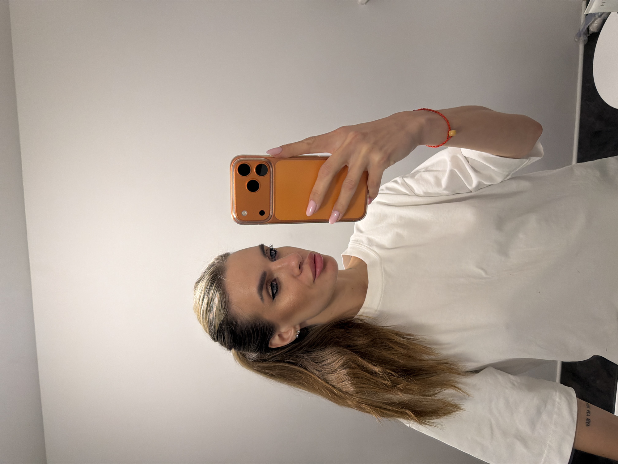 A woman is depicted taking a mirror selfie indoors. She has long, multi-toned blonde and brown hair styled in a high ponytail. Her face features prominent eye makeup, including winged eyeliner, and contoured cheeks, with light pink lipstick. She wears small, clear stud earrings. Her attire consists of a loose-fitting white short-sleeved t-shirt. A tattoo of inverted text, partially reading "YAW YM," is visible on her left forearm. On her right wrist, she wears a red string bracelet with a single round, yellowish bead.
She holds an orange-cased smartphone, equipped with three visible rear camera lenses, in her right hand. Her fingernails are painted a light pink. The woman's gaze is directed towards the phone screen within the reflection. The immediate environment consists of a plain, light-colored wall, likely white or off-white. A small portion of a white surface, possibly a counter or shelf, is visible in the lower right, with indistinct white and metallic objects resting upon it. The exact location within Berlin, Germany, cannot be determined from the visual information provided.