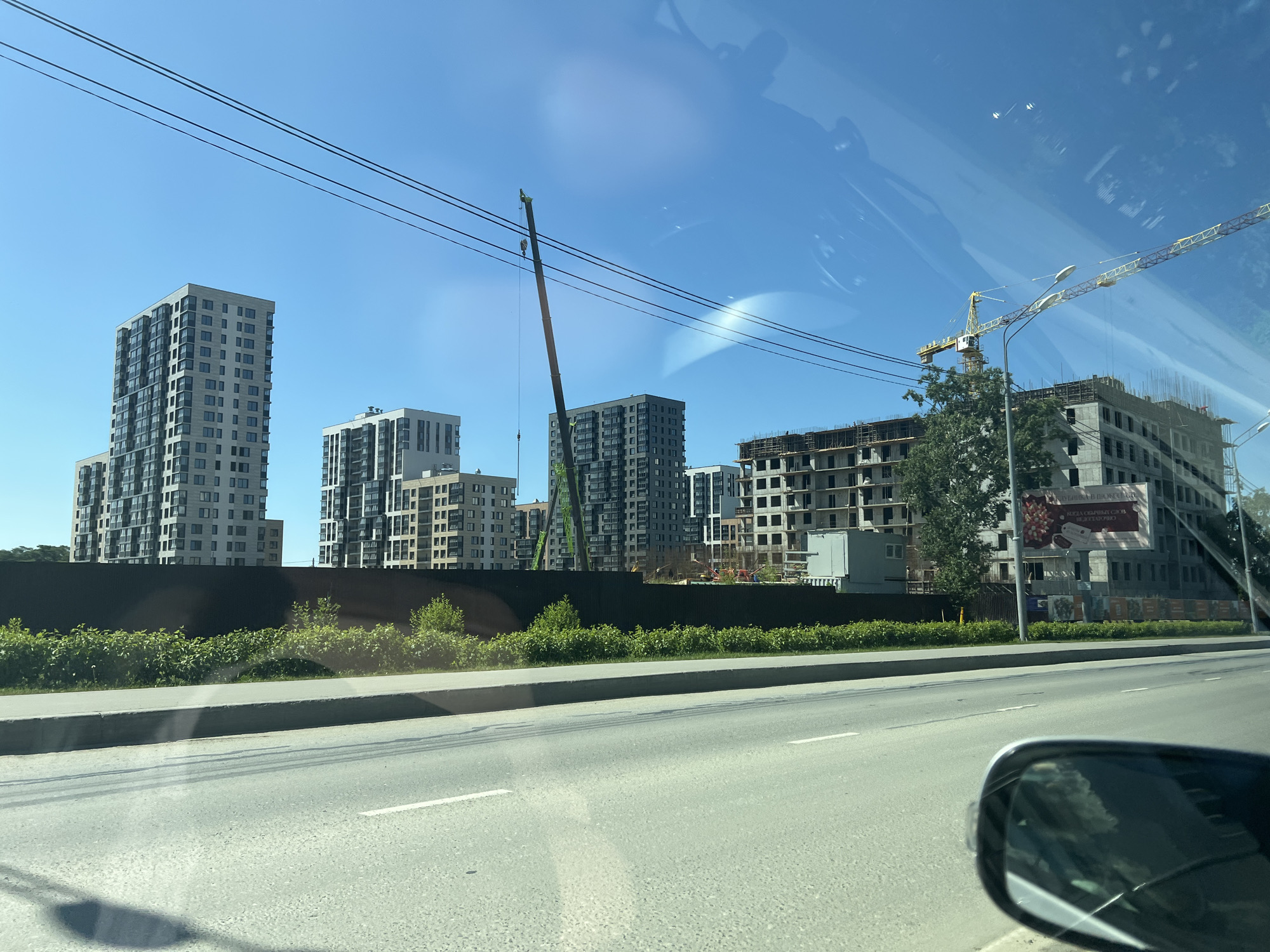 The image shows a street view in Yuzhno-Sakhalinsk, Russia, on a clear day. In the background, several multi-story buildings are under construction, with visible concrete structures, scaffolding, and cranes. Some completed apartment buildings are also present. A dark fence lines the perimeter of the construction site, with a row of green bushes in front of it. The foreground features a paved road with white lane markings and a sidewalk. A car's side mirror is visible in the lower right corner, reflecting the surrounding environment. Power lines are strung across the sky. The scene depicts an urban area with ongoing development.