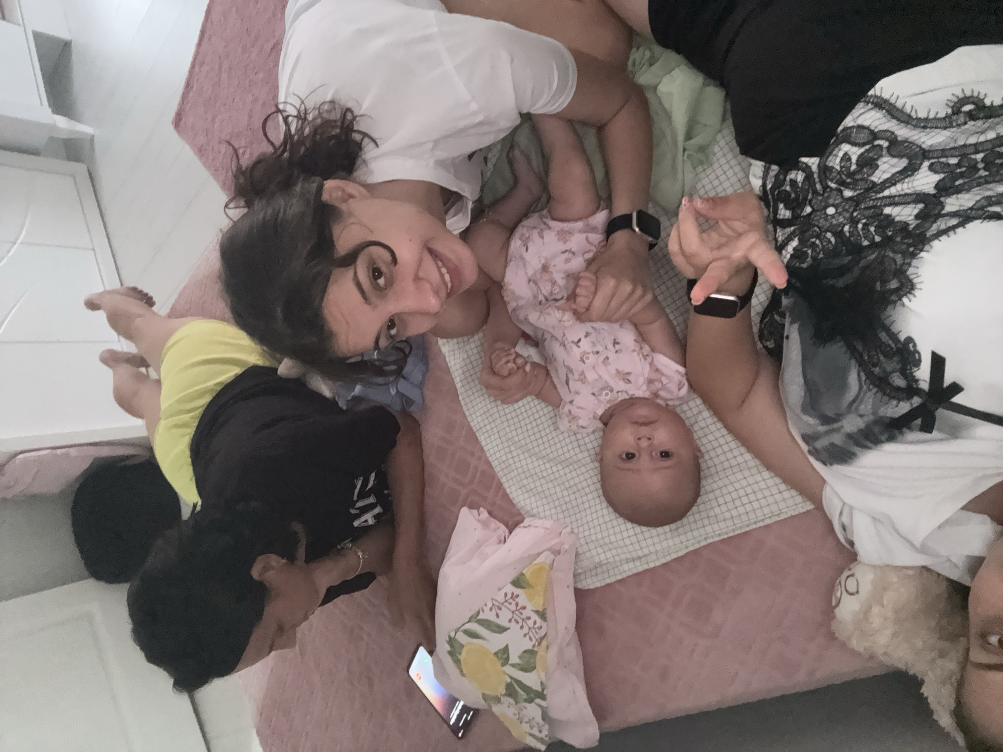The image depicts an indoor scene on a bed, likely in a home located in Stamboliyski, Bulgaria. Three individuals are visible: an adult woman with dark hair lying on her side, propped up on her arm, looking down; a baby lying on its back on a checkered blanket, wearing a pink floral outfit; and another adult woman with dark, curly hair in a ponytail, leaning over the baby and smiling at the camera. The background includes a pink textured bedspread, a white dresser, and a pink rug. Objects present include a smartphone on the bed, a decorative pillow with lemons, and a stuffed animal. The women are dressed casually, one in a black t-shirt and the other in a white t-shirt. The overall impression is of a relaxed, familial moment.