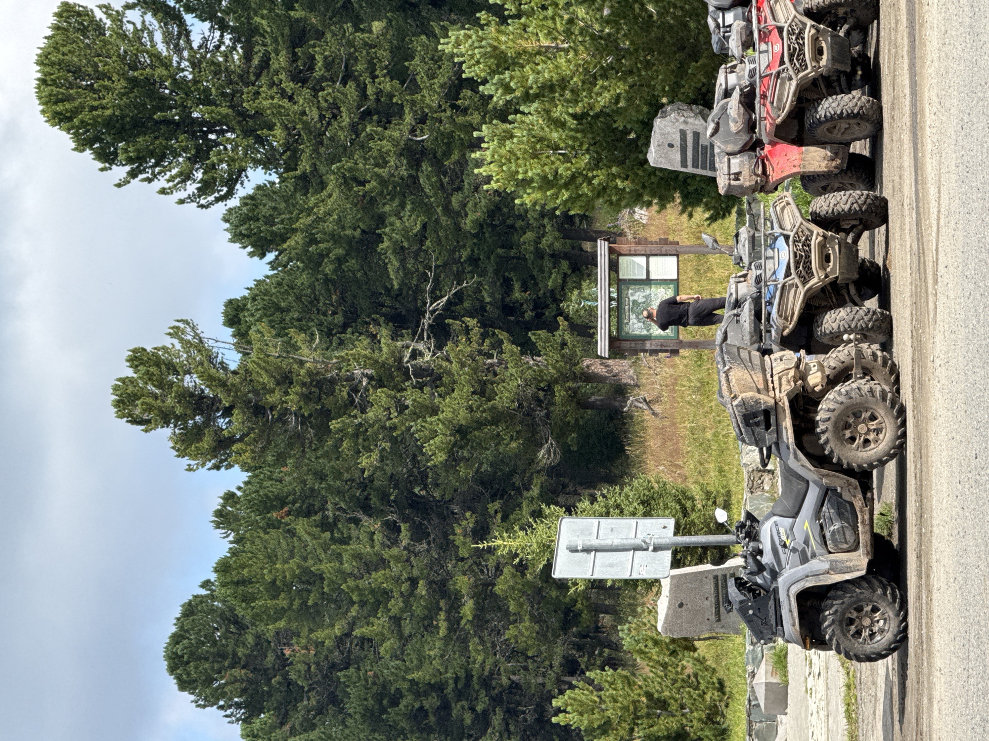 The image captures a scene at T/B Seminskii Pereval, Russia, with several ATVs parked on a dirt road in the foreground. The ATVs are various colors, including gray, blue, and red, and appear to be used for off-road excursions, as indicated by the mud on their tires and bodies. In the background, a person is standing near a wooden information board, seemingly reading the displayed content. Behind the information board and person, a dense forest of tall pine trees stretches across the landscape, under a partly cloudy sky. The environment suggests a natural, mountainous area, typical of a mountain pass.