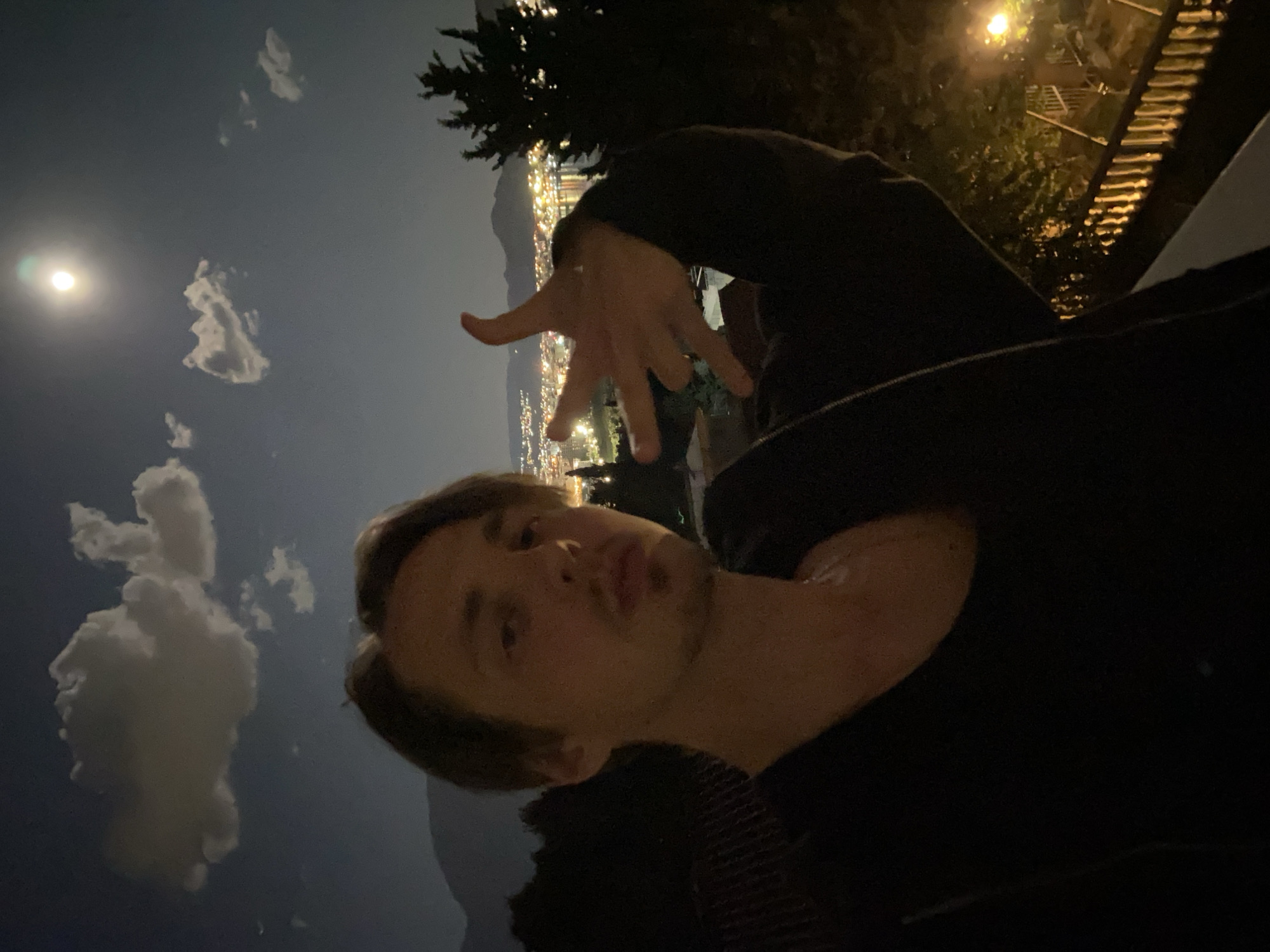 A selfie of a young man with light skin and brown hair. He's wearing a black shirt and is posed with his hand outstretched towards the viewer. The background is a nighttime cityscape with visible city lights, and a bright full moon is visible in the sky. Some clouds are present. In the upper part of the background, there is what appears to be a tree and a street light. The location is likely Šušanj, Montenegro, based on the coastal cityscape visible in the background.