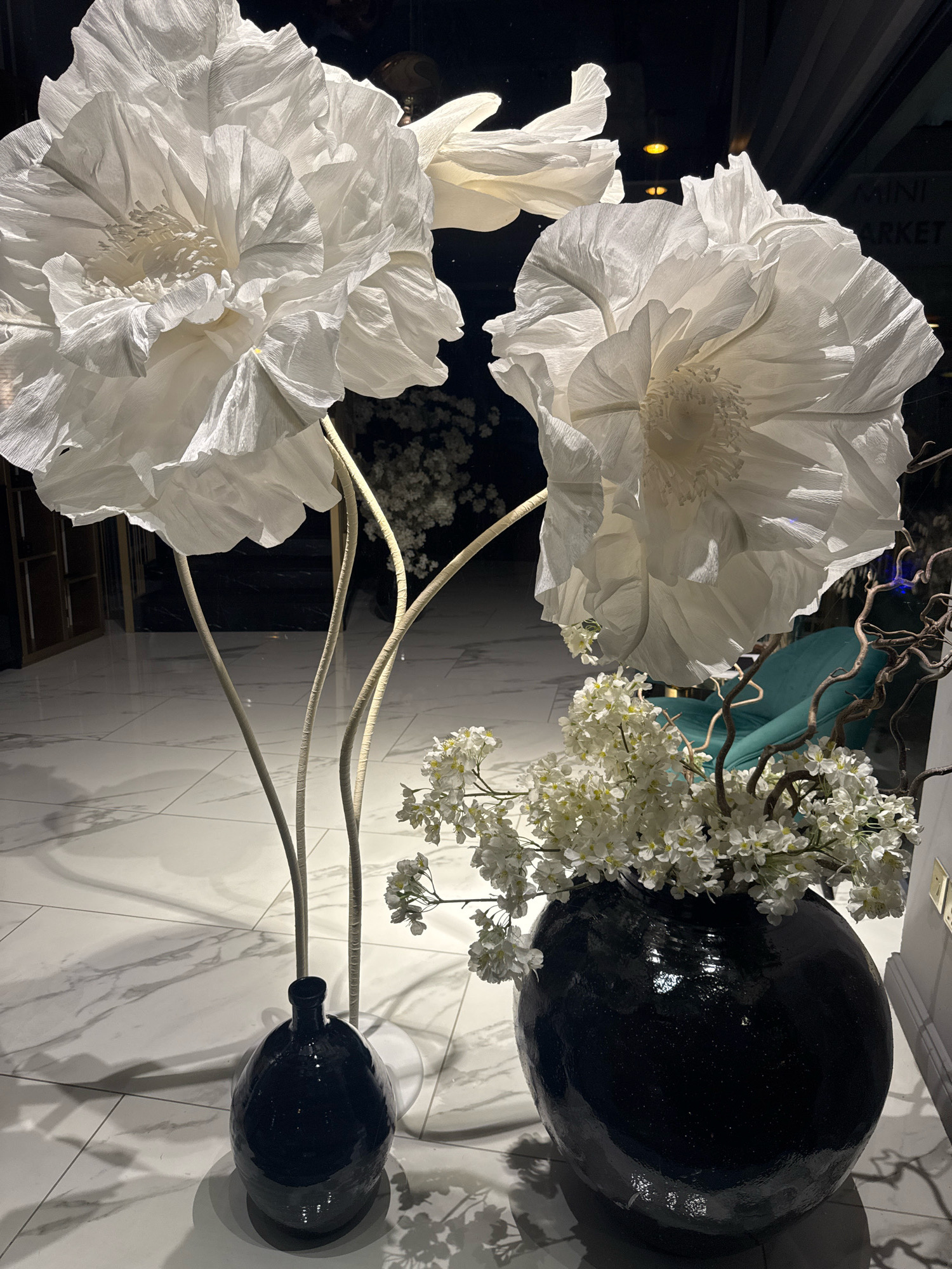 The image displays a dimly lit interior scene featuring oversized white paper flowers and a black vase filled with white flowers. The large paper flowers are the primary focus, with intricate details in their petals. To the right, a black, glossy vase holds a bouquet of smaller white flowers and dried branches. In the background, a teal armchair is visible, and a sign that reads "MINI MARKET" can be seen. The floor is a polished marble tile. Based on the context provided about Limassol, Cyprus, this scene could be located within a shop or a public space in the city, possibly a flower shop, a decoration store, or an event venue. There are no people visible in the image.