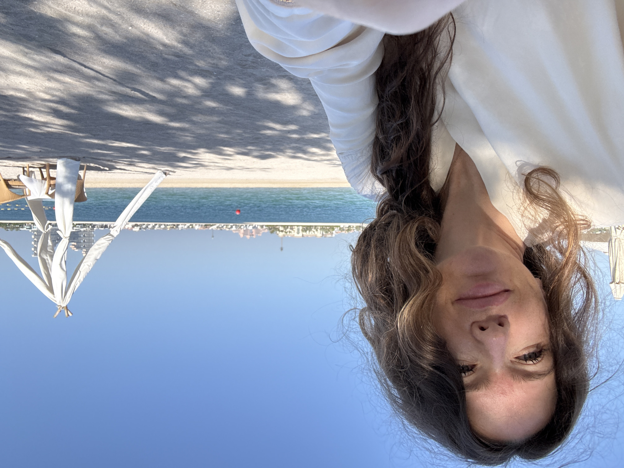 A selfie is taken by a woman with long, wavy brown hair, facing the camera. She is wearing a white top. Her face is partially lit by the sun. In the background, a beach scene unfolds with a calm blue sea and a clear sky. A white canopy tent is erected on the sand, with two wicker chairs placed nearby. Distant buildings are visible across the water, and a red buoy floats in the sea. The overall impression is a sunny day at a coastal resort. The buildings in the distance are consistent with the architecture found on the Palm Jumeirah in Dubai, United Arab Emirates, suggesting the location is likely this artificial island.