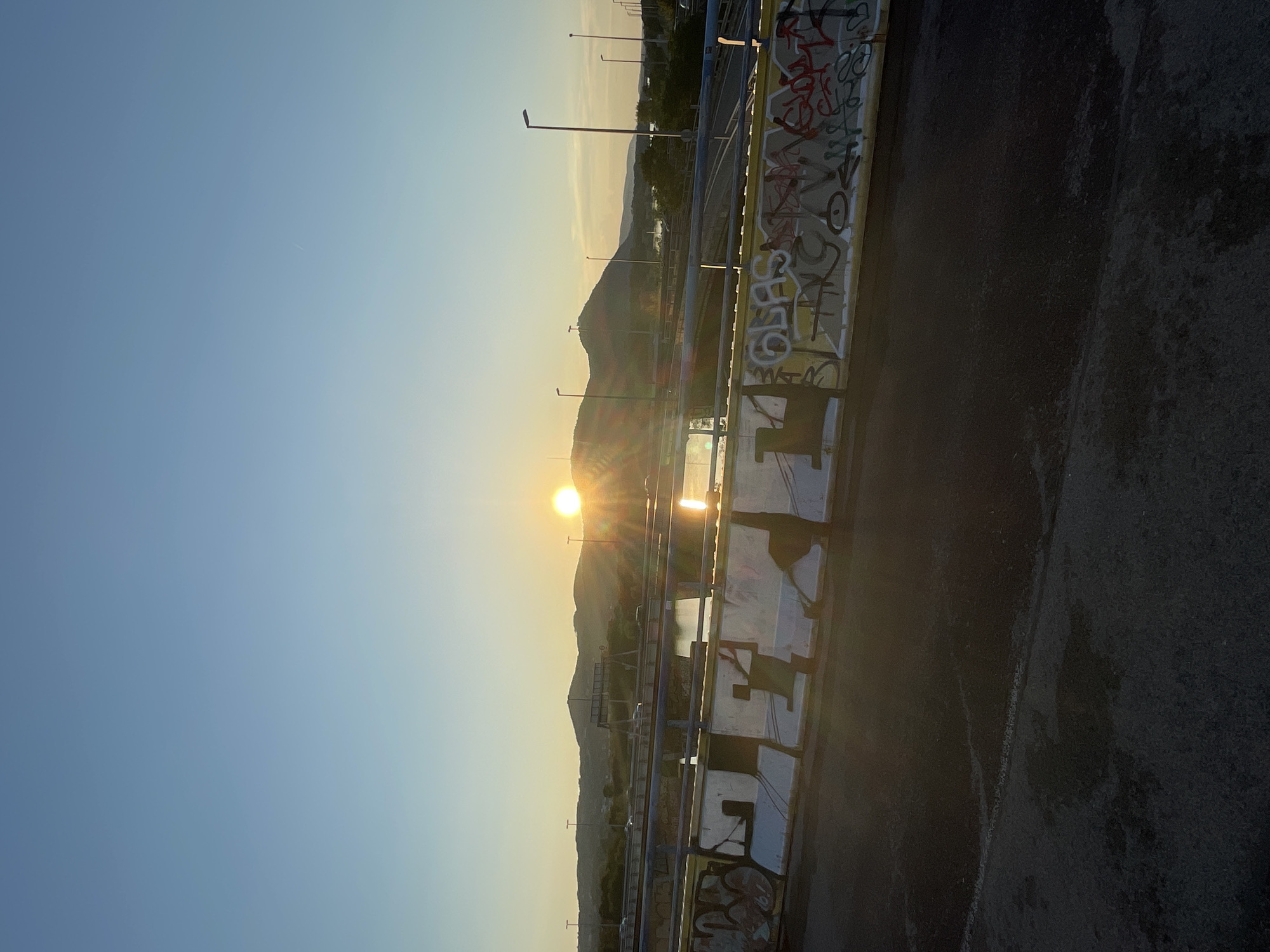This is a landscape photograph taken at sunset, likely from a bridge, overlooking a body of water with a distant cityscape and hills. The sun is setting behind the hills, casting a warm glow and creating a reflection on the water. The foreground of the image features a concrete barrier covered in graffiti, with a roadway visible behind it. The sky is clear and blue, with a gradient towards a lighter hue near the horizon. There are streetlights visible on the bridge and in the distance, along with a few cars driving on the road. The overall scene suggests an urban environment at dusk. This image is not from Vienna, Austria.