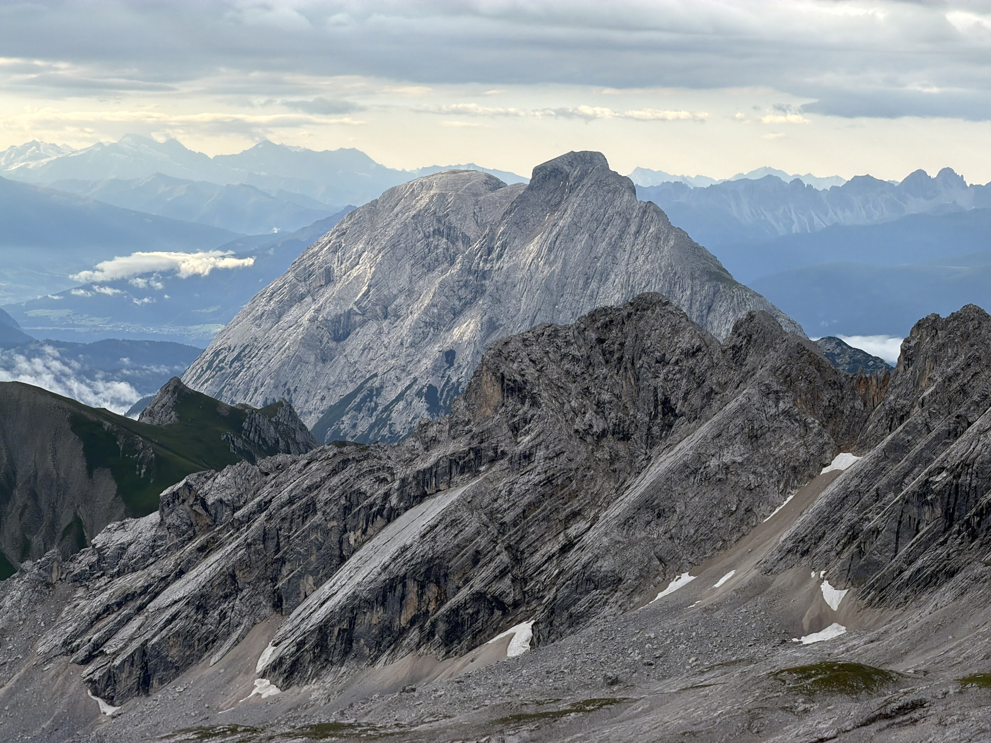 The image depicts a majestic mountain range, characterized by rugged, rocky slopes and layered stratifications. In the foreground, steep inclines covered in scree and scattered patches of snow dominate the landscape. The rock faces are textured, showing fissures and variations in color from light gray to darker shades. Further into the background, a prominent, craggy mountain peak rises, its summit appearing to have a cross or antenna. Beyond this central peak, a series of progressively hazier mountain ranges stretch towards the horizon, fading into a soft blue hue. The sky above is partially covered with clouds, some appearing wispy and light, while others are denser and darker, suggesting a dynamic weather system. The overall scene conveys a sense of vastness and natural grandeur. While the image shows a mountainous landscape, it is not possible to definitively pinpoint a specific location within Garmisch-Partenkirchen, Germany, based solely on the visual information provided.