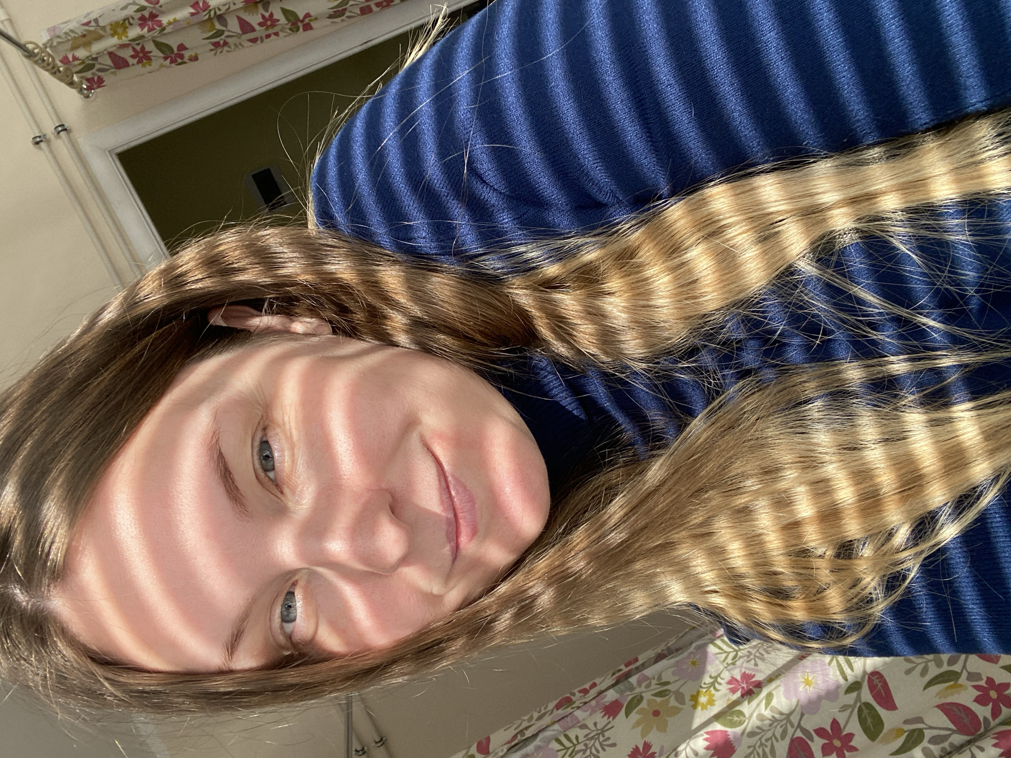 An indoor, brightly lit portrait of a young woman, likely taken in Noviy, Russia, presented from a high-angle perspective. The woman's head is tilted to the left, and she has a gentle, closed-mouth smile. Her light blue eyes look directly towards the viewer.
A striking feature is the strong, parallel stripes of golden sunlight and shadow cast across her face, long, light brown hair, and the dark blue, possibly knitted or ribbed, garment she is wearing. These shadows suggest bright natural light filtering through window blinds, indicating a sunny morning or late afternoon. Her hair, which shows blonde highlights where the sun hits, flows across her shoulder and onto a floral-patterned fabric, possibly bedding or a blanket. This fabric features an array of pink, green, and yellow flowers on a light background.
In the upper left background, a light-colored wall and part of a doorframe are visible. The overall mood is serene and pleasant due to her expression and the warm sunlight. No text is visible in the image.